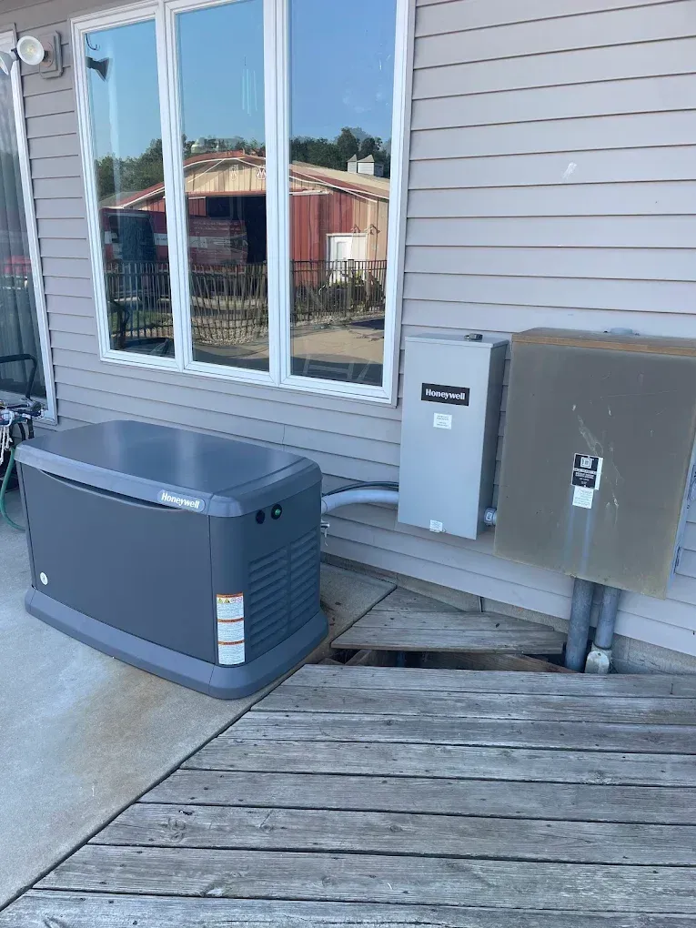 A gray generator, switch box, and electrical panel are next to a house with large windows.