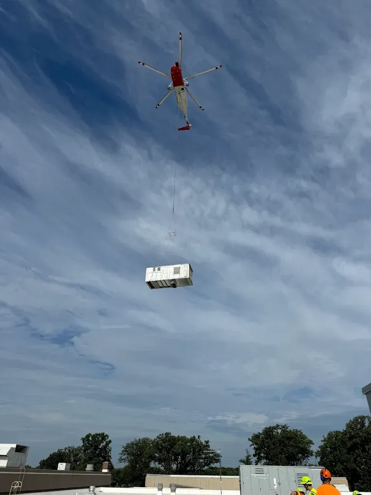 Helicopter lifting a white building module, suspended in air. Blue sky, clouds, and a person in a safety vest below.