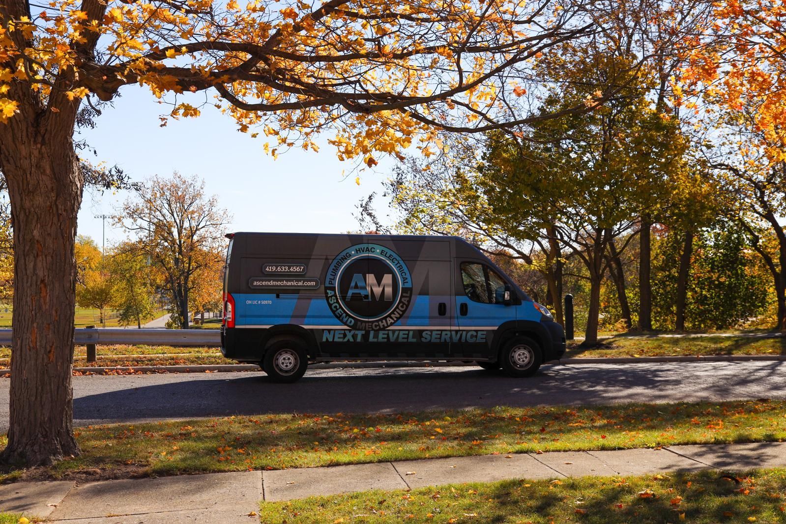 Blue work van with logo parked on a road, fall foliage, sunny day.