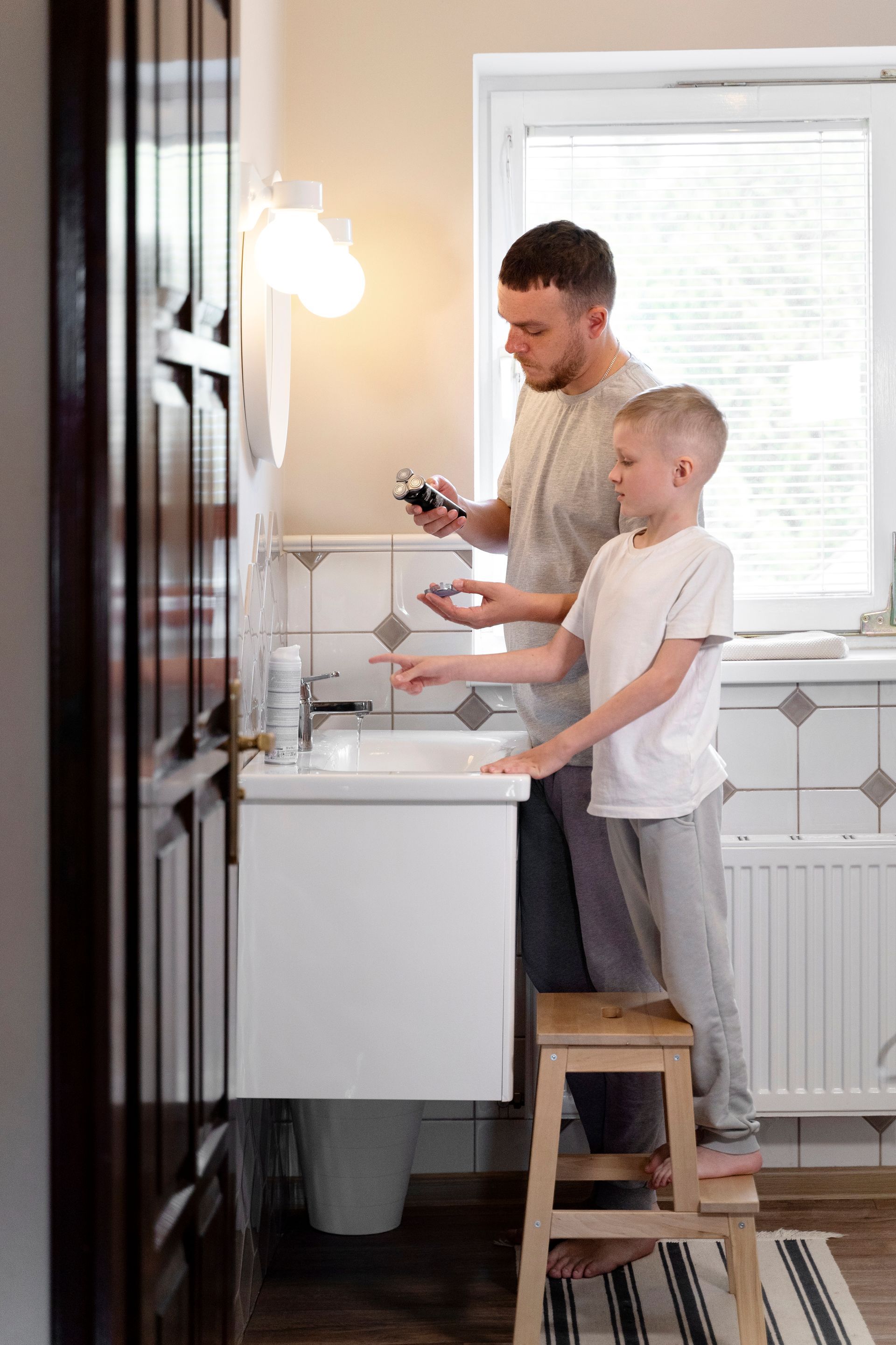 Father and child washing hands in a bathroom. Child on a step stool, pointing at the faucet.