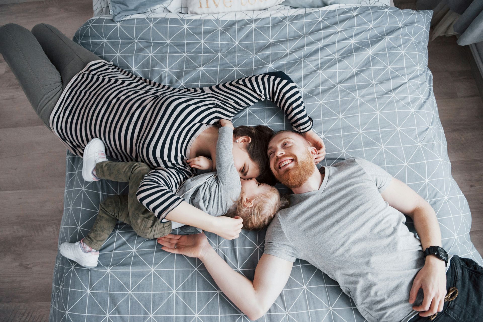Family of three cuddling on a bed with a gray patterned comforter, smiling.