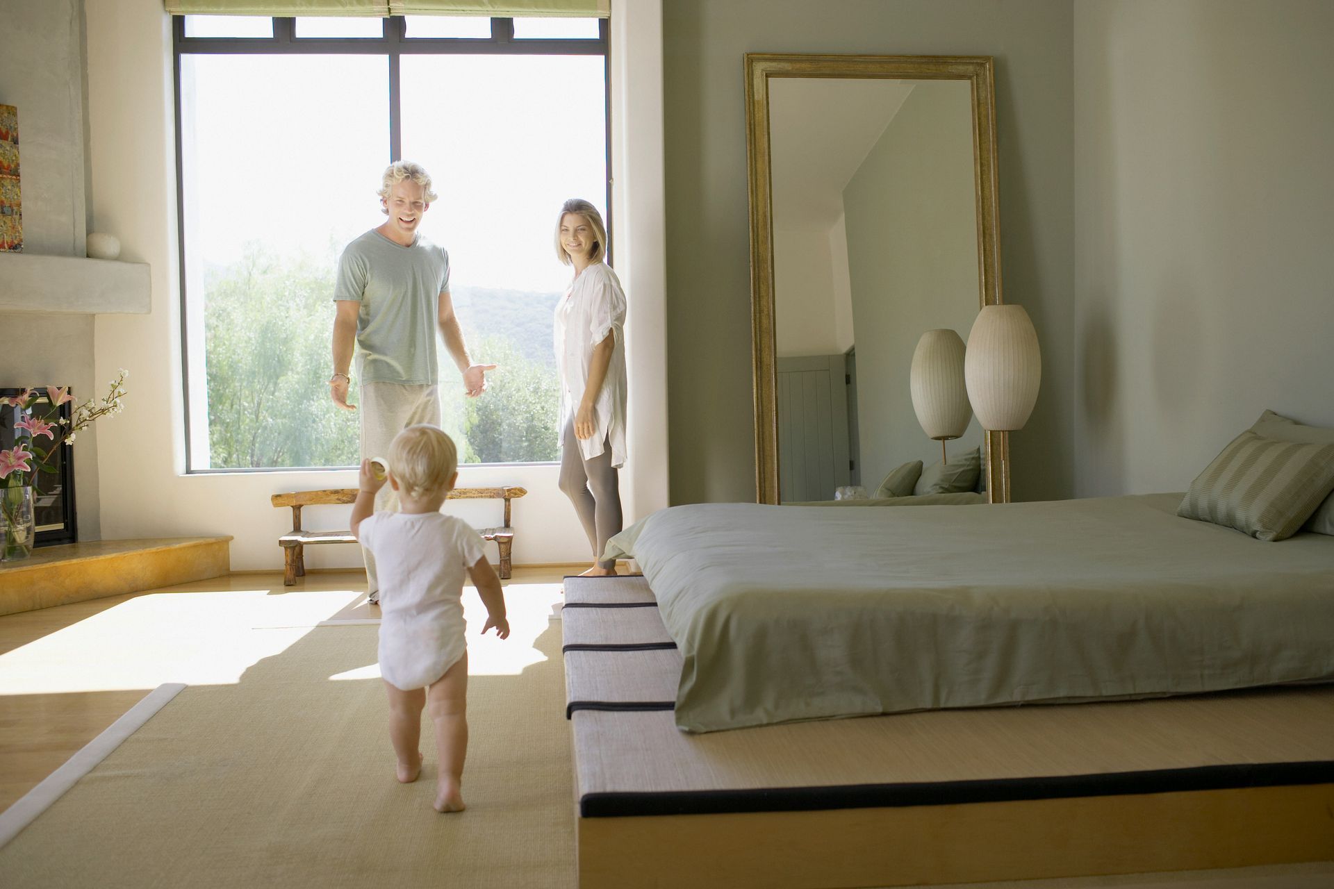 Child walking toward parents in bedroom with large mirror.