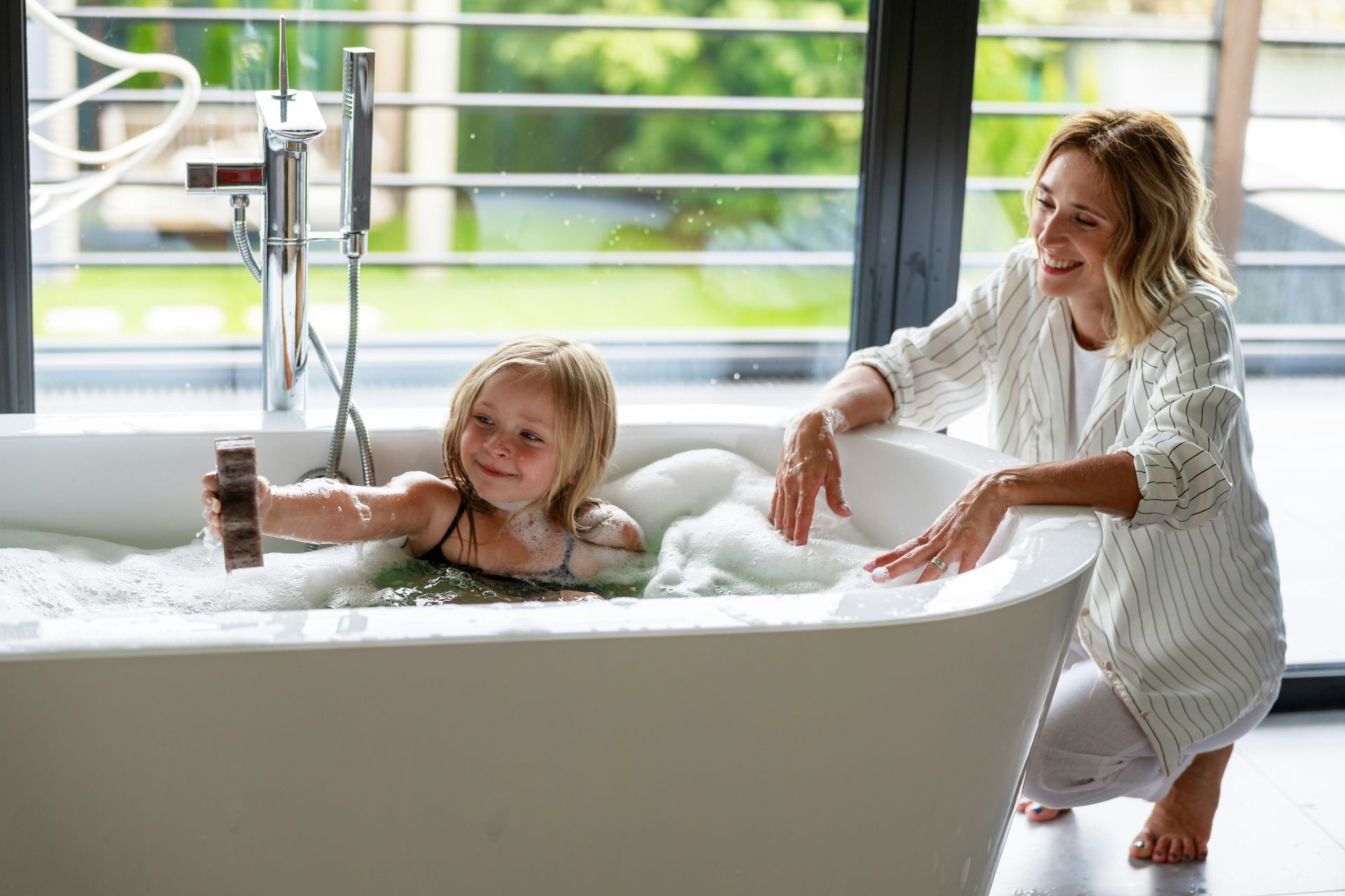 A woman watches a child playing in a bathtub filled with bubbles; large windows in background.