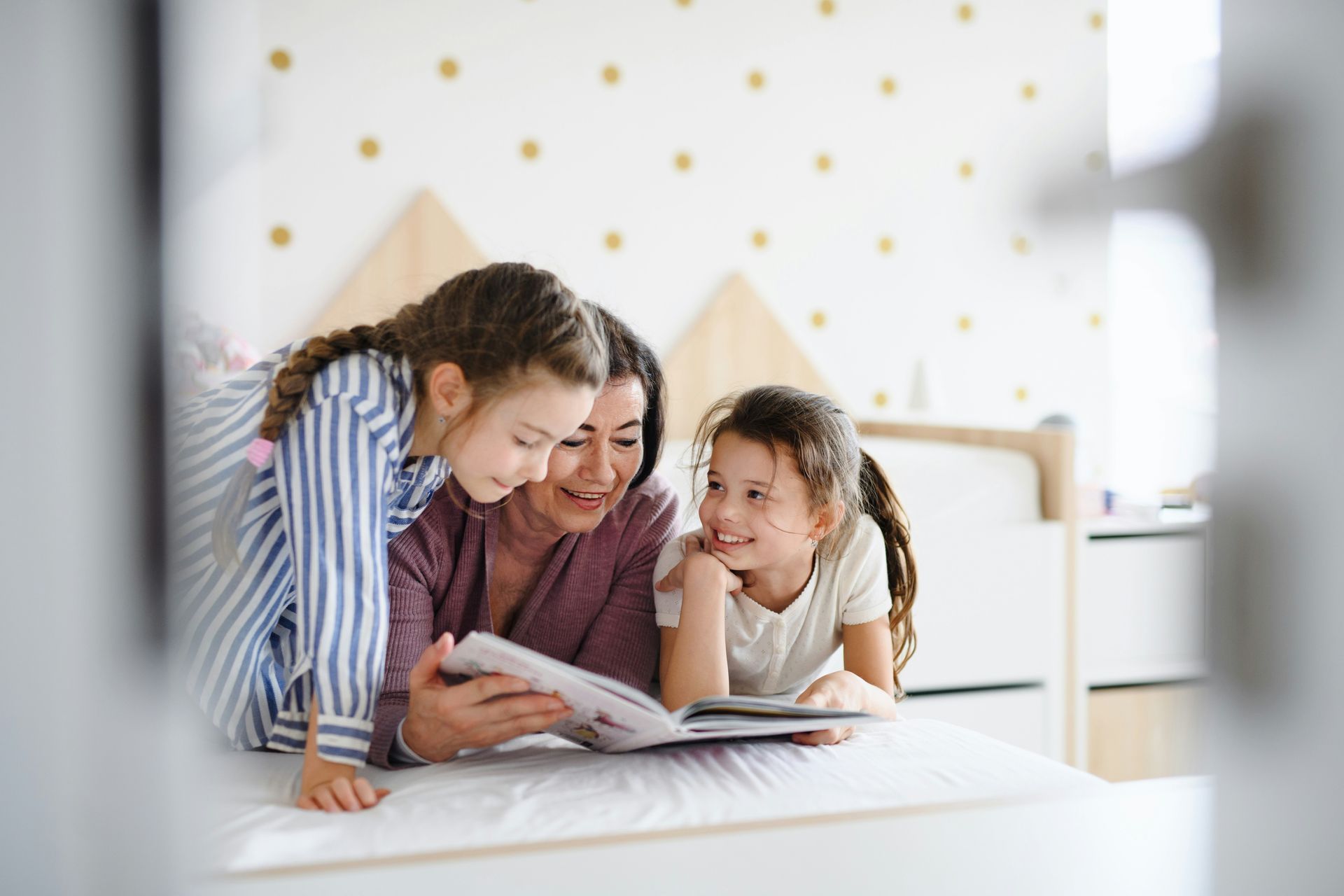 Woman and two children read a book together on a bed; the children look at the book.