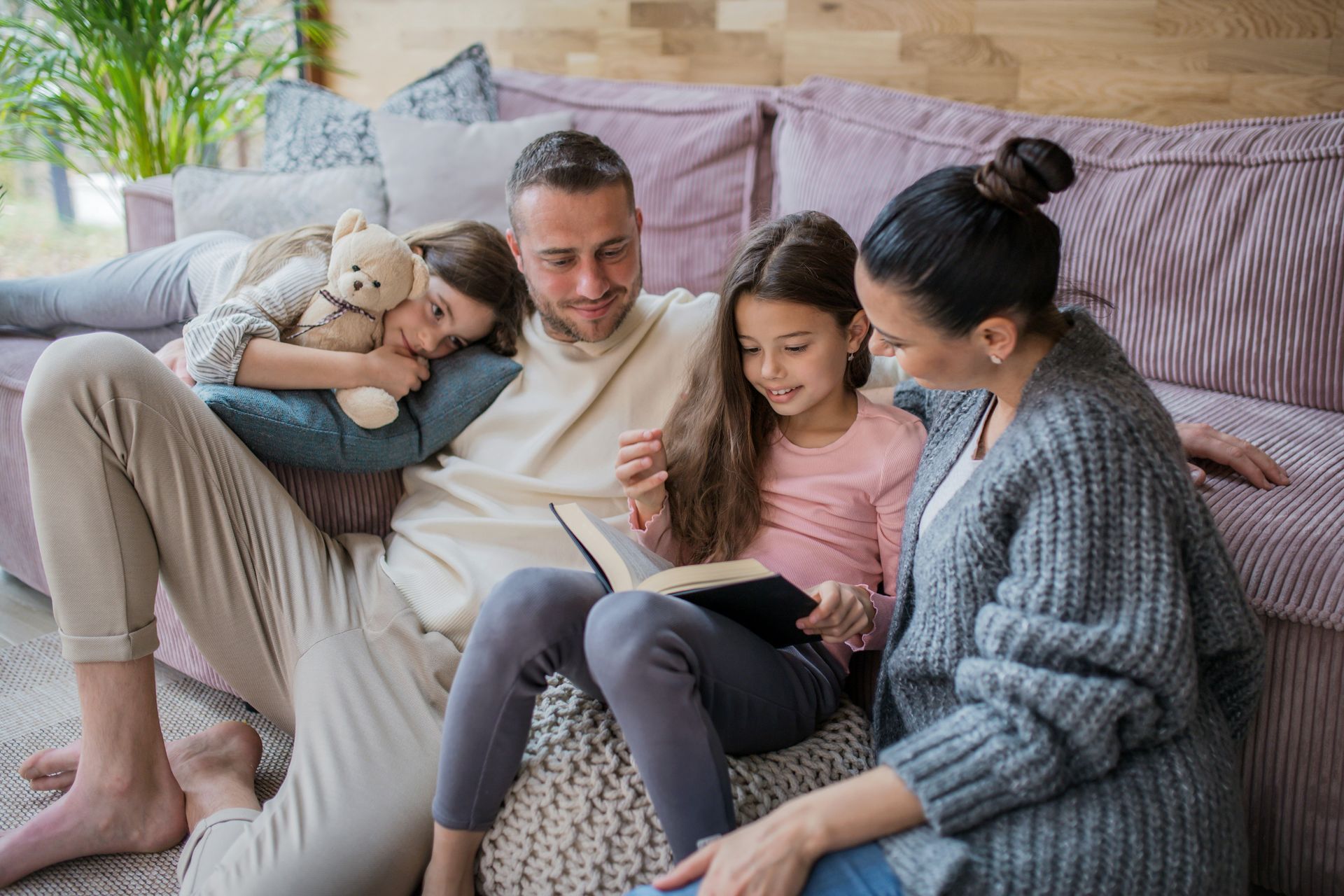 Family of four sitting on floor and couch reading a book together in a living room.
