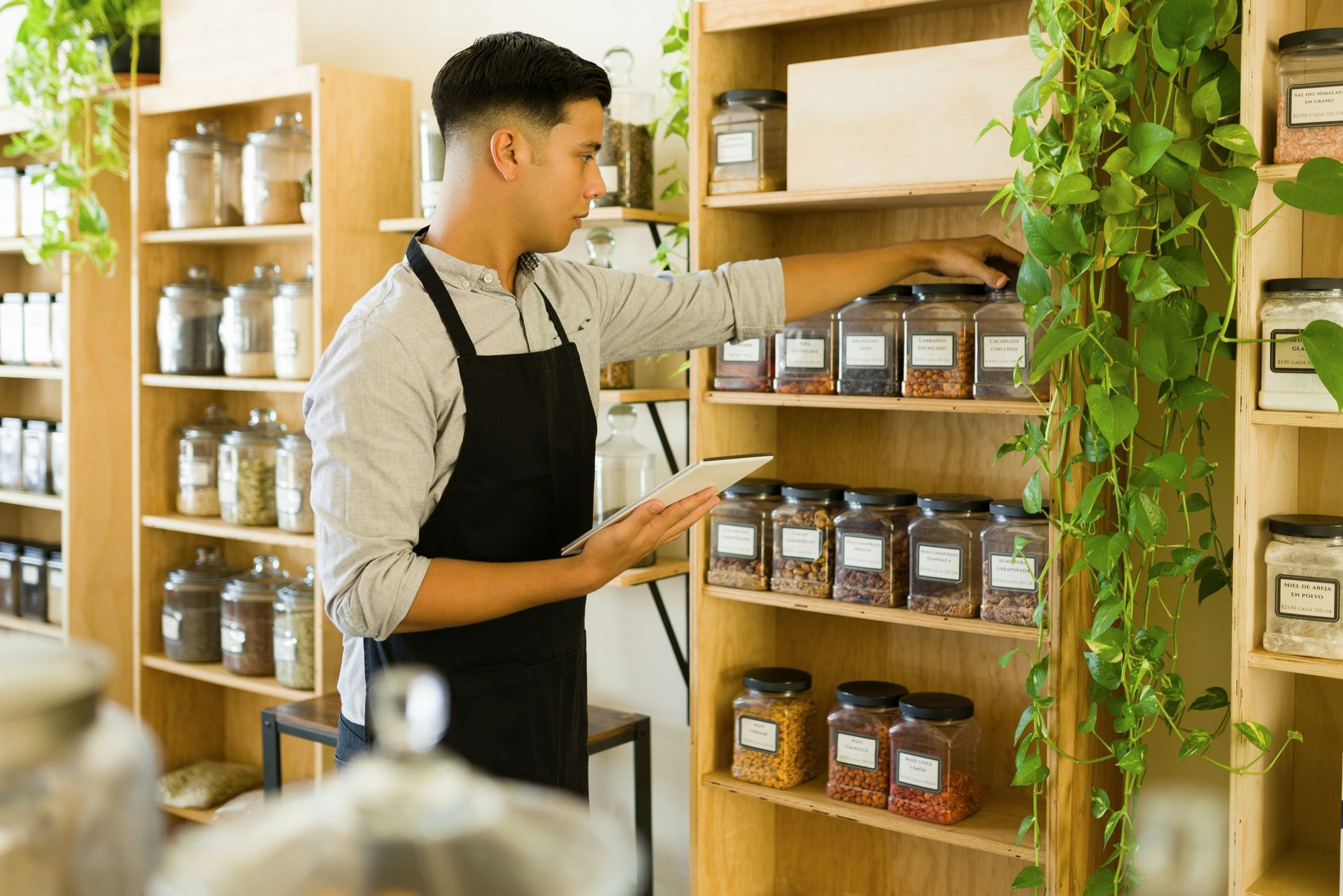 A person wearing an apron checks inventory of jarred goods on wooden shelves in a store.