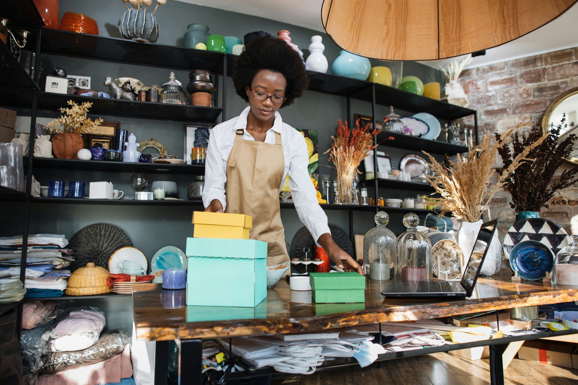 Woman in apron arranging gift boxes on a counter in a shop with shelves of items.