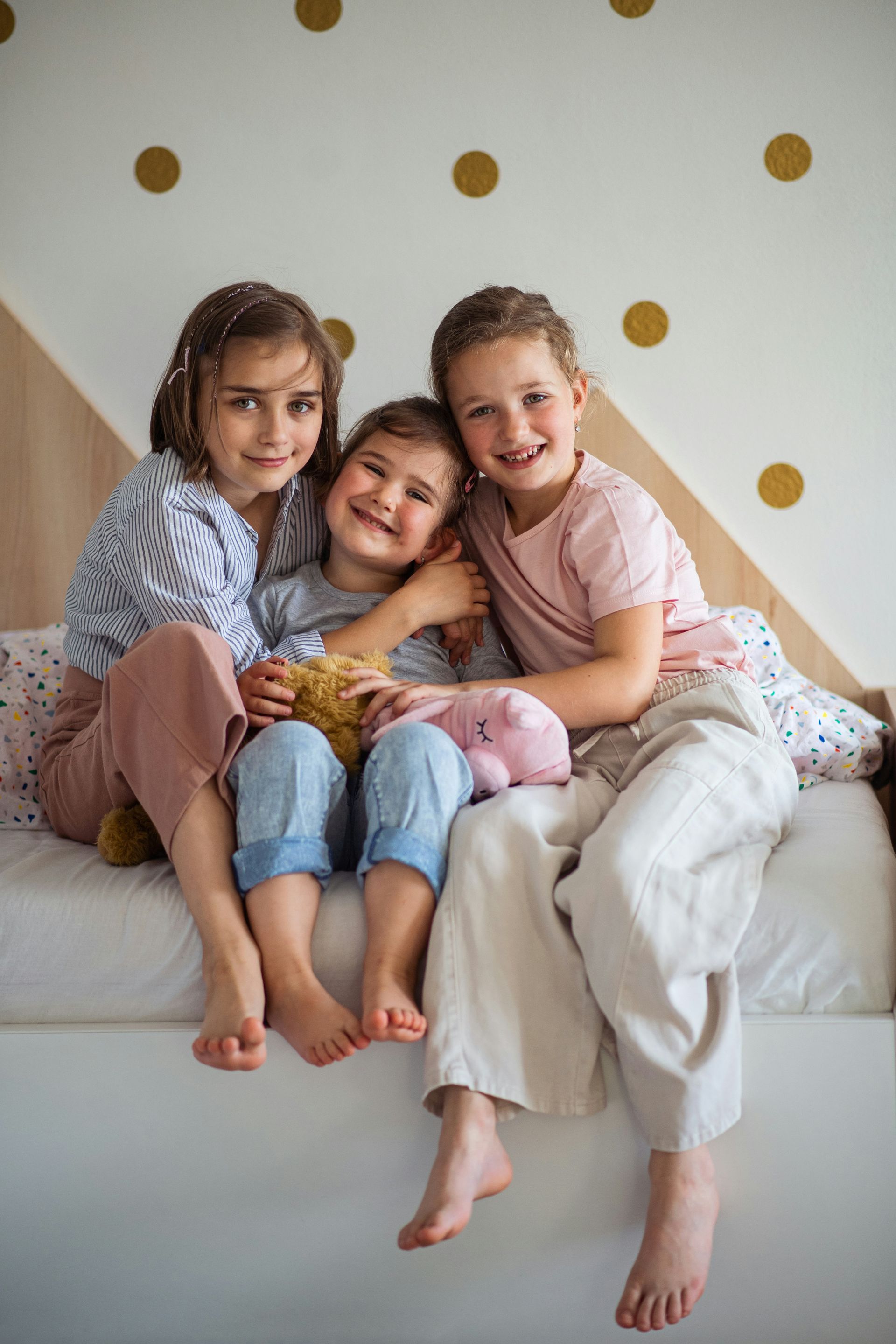 Three children smiling, sitting close together on a bed; bedroom setting with gold polka dots on wall.