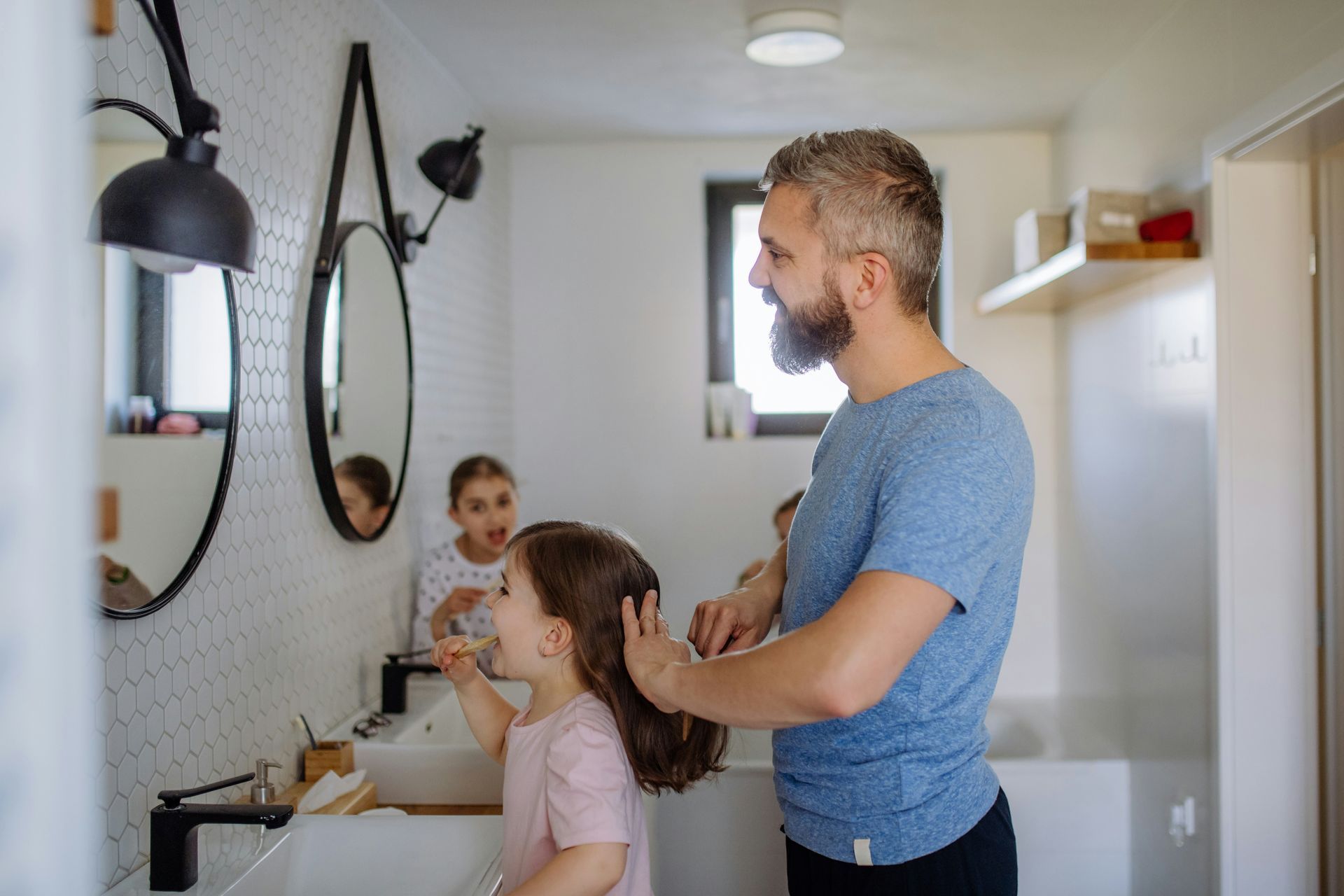 Man combing girl's hair in a bathroom. Two children in background at the sink, mirrors on the wall.
