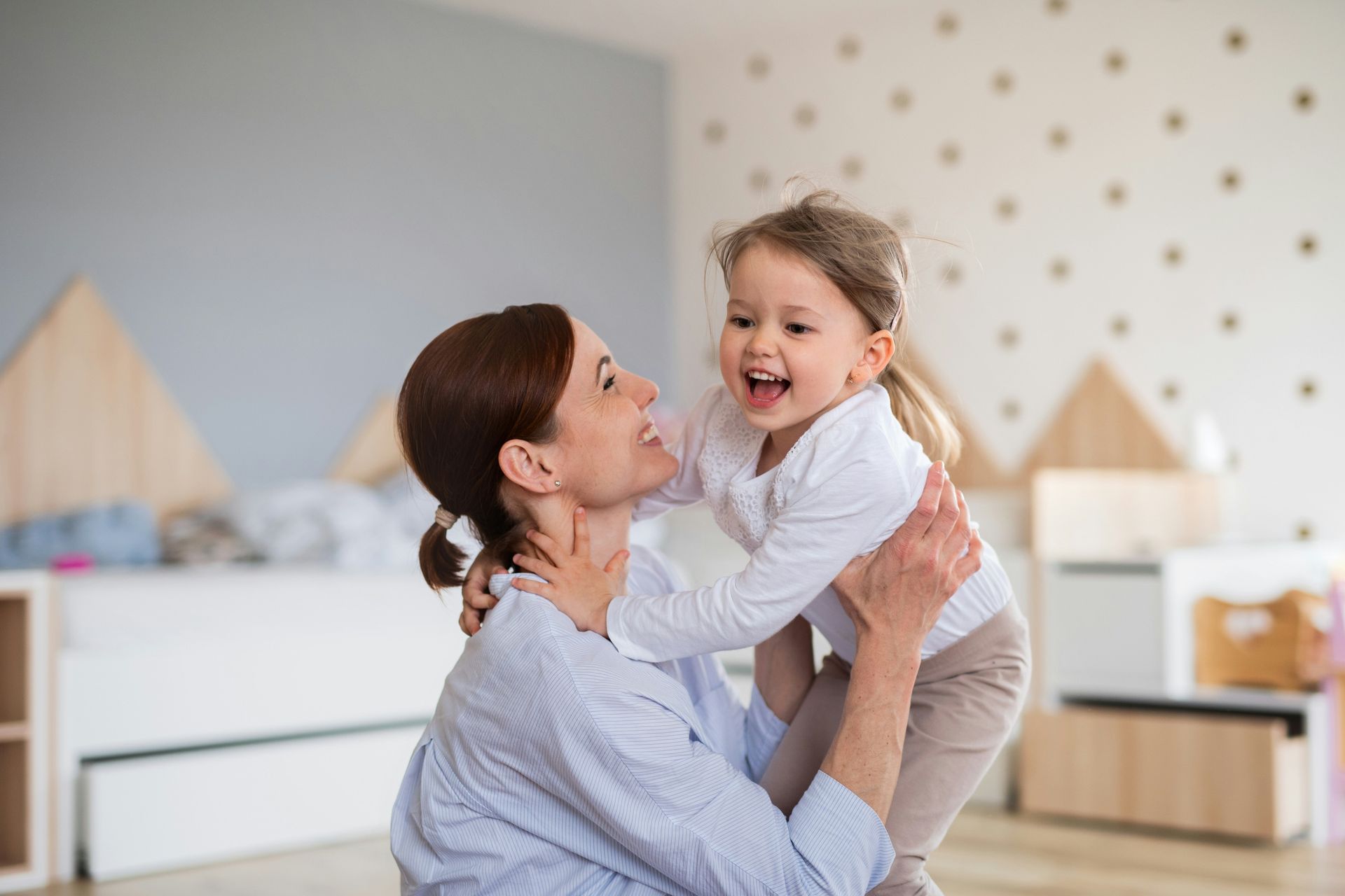 Woman holding a laughing child in a bedroom; light blue walls and decor.