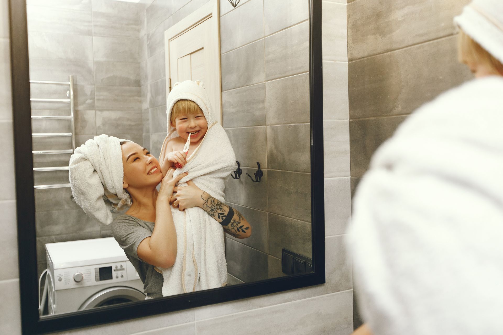 Woman holds child wrapped in a towel, smiling in bathroom mirror.