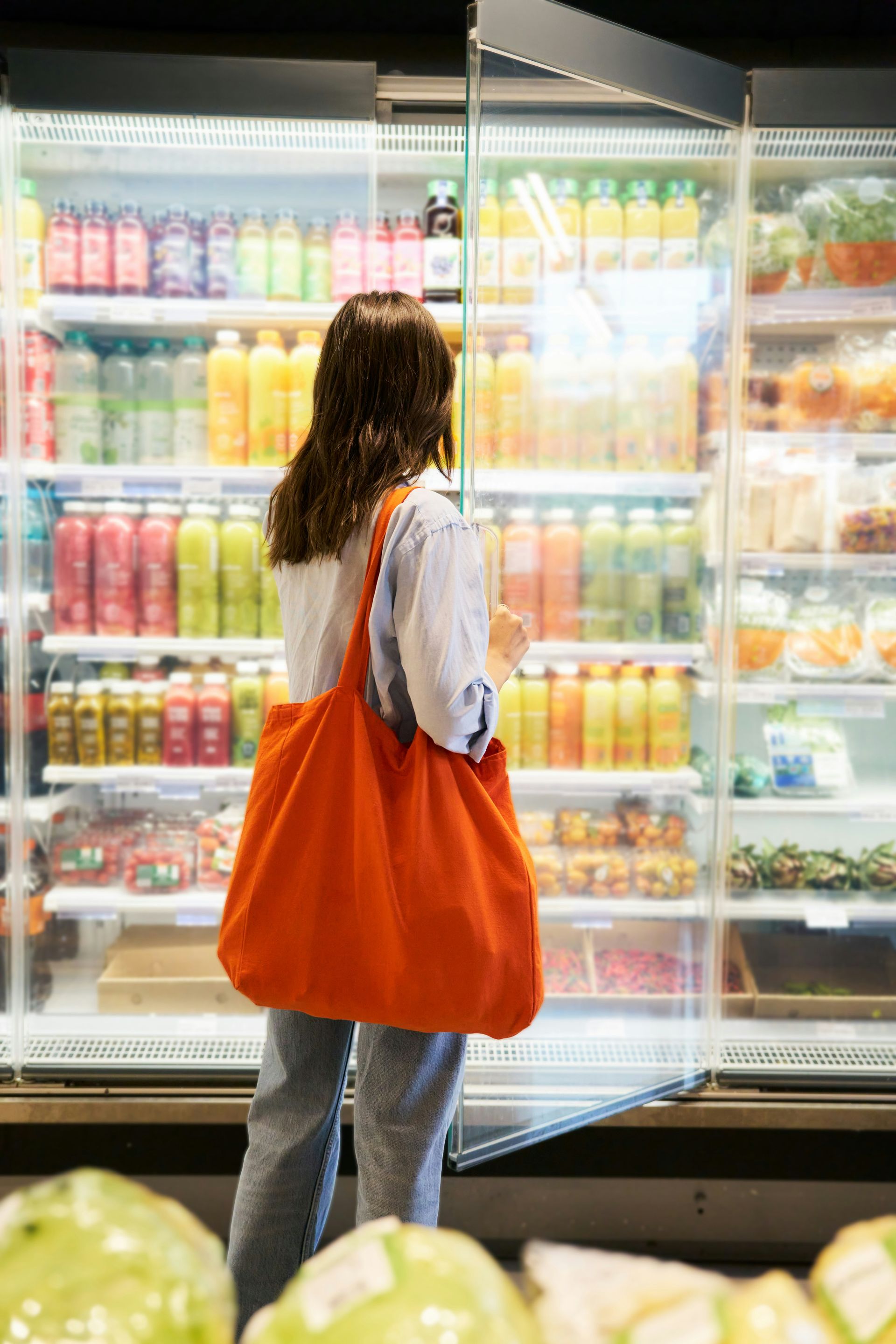 Woman with orange tote bag browsing refrigerated food display at a grocery store.
