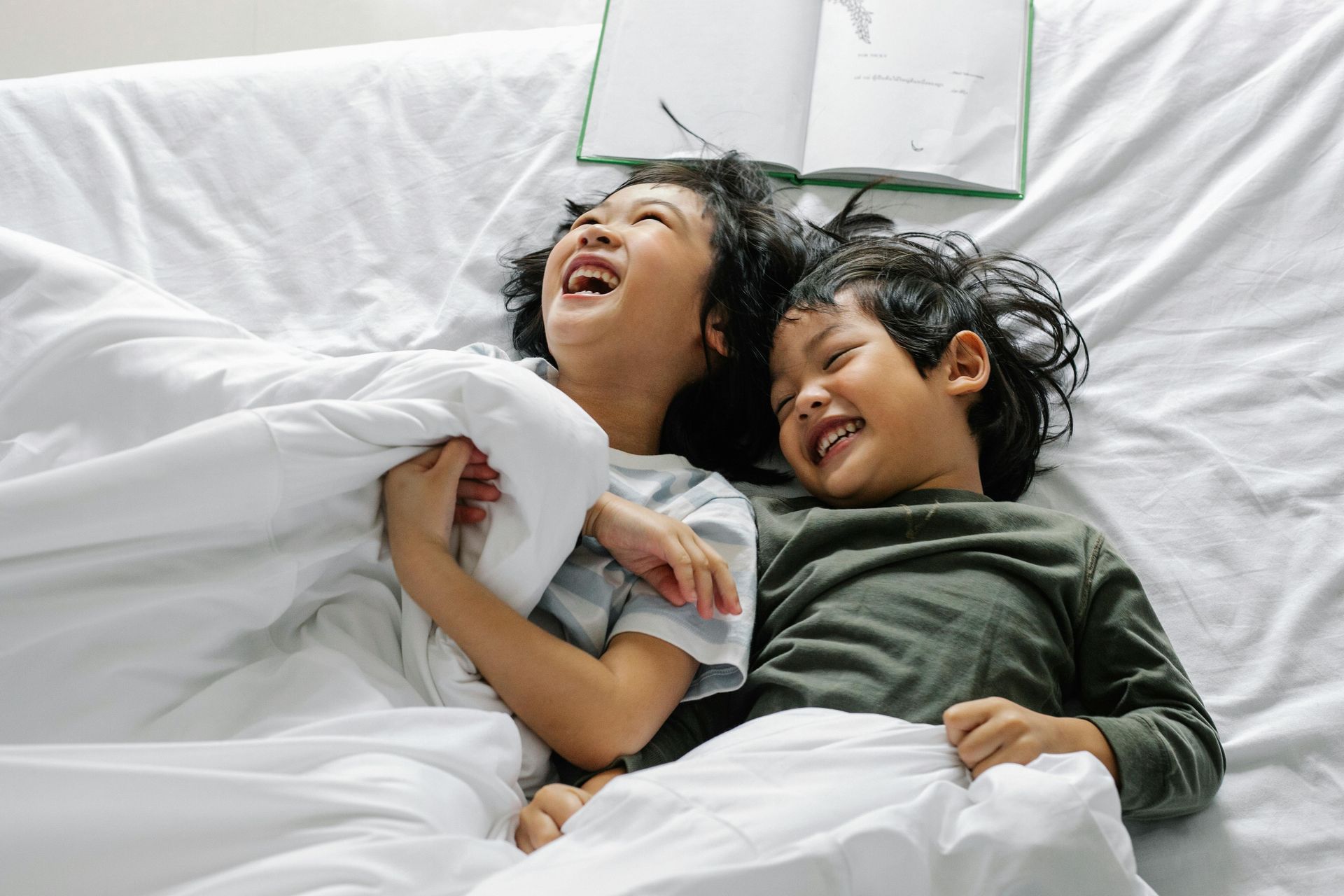 Two children laughing and playing on a bed, covered by a white blanket, with an open book nearby.