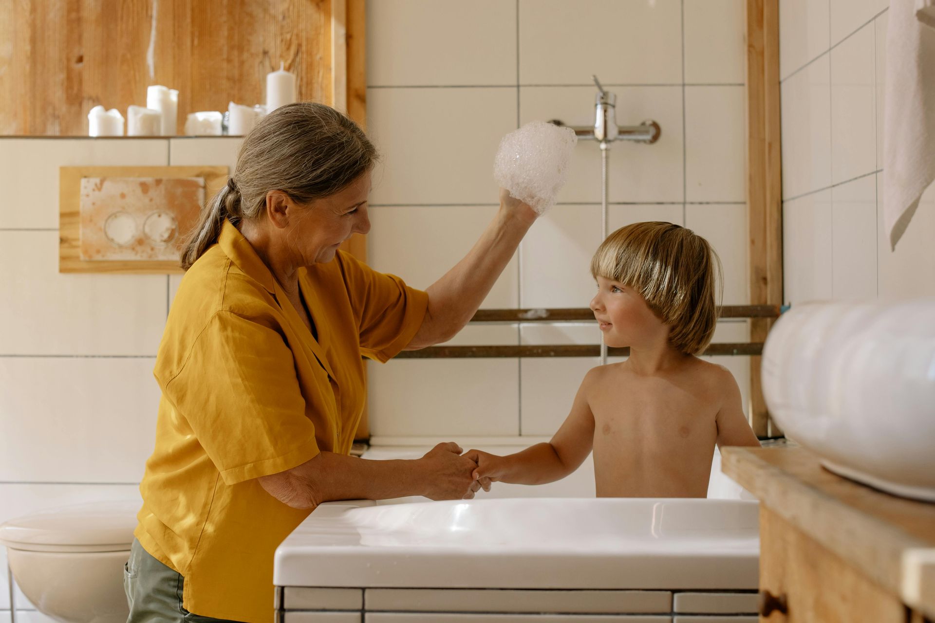 Woman in yellow shirt, bathes child in a white tub, playing with soap suds in a bright bathroom.