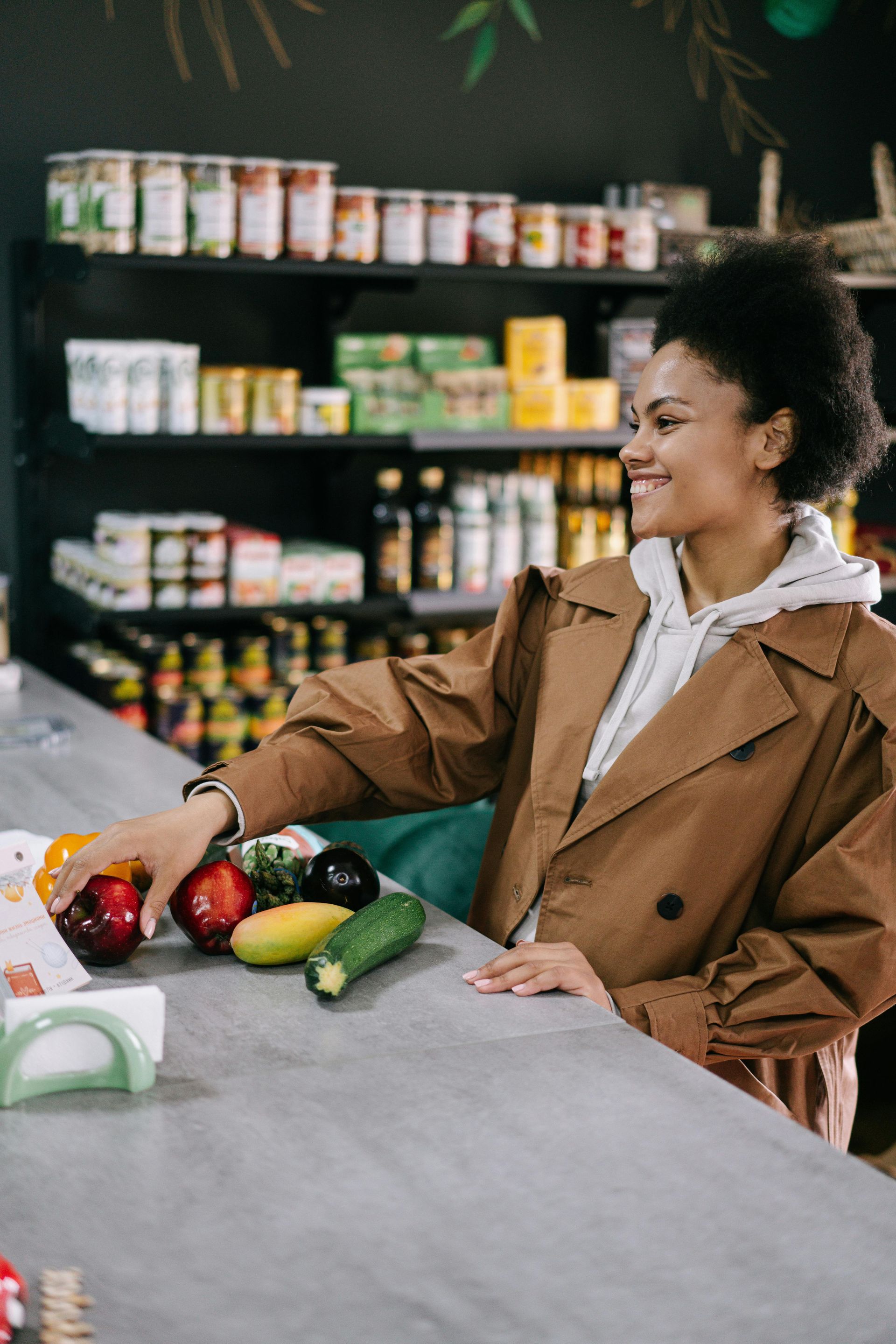 Woman smiles at fresh produce on a store counter. Shelves stocked with food are behind her.