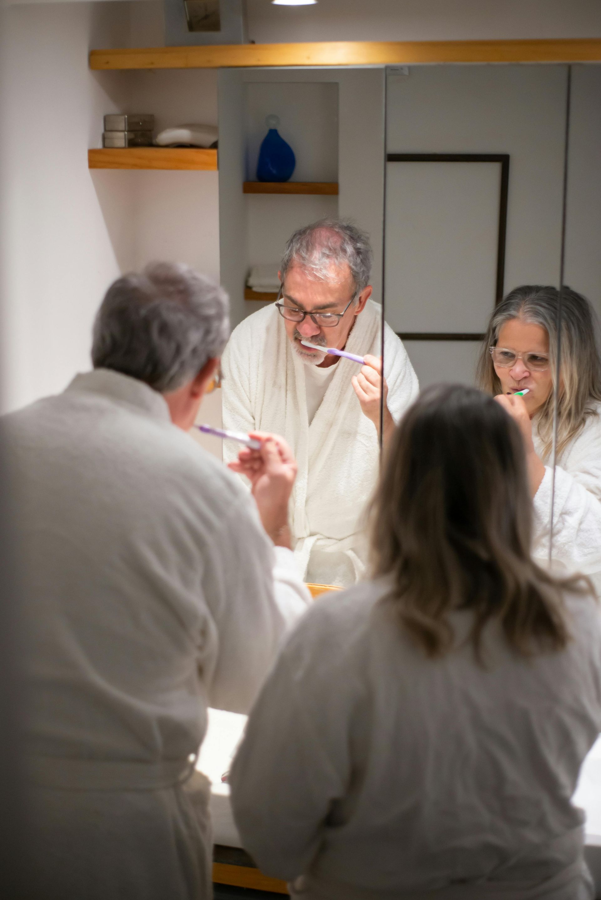 Three people in white robes brushing their teeth in a bathroom, reflected in a mirror.