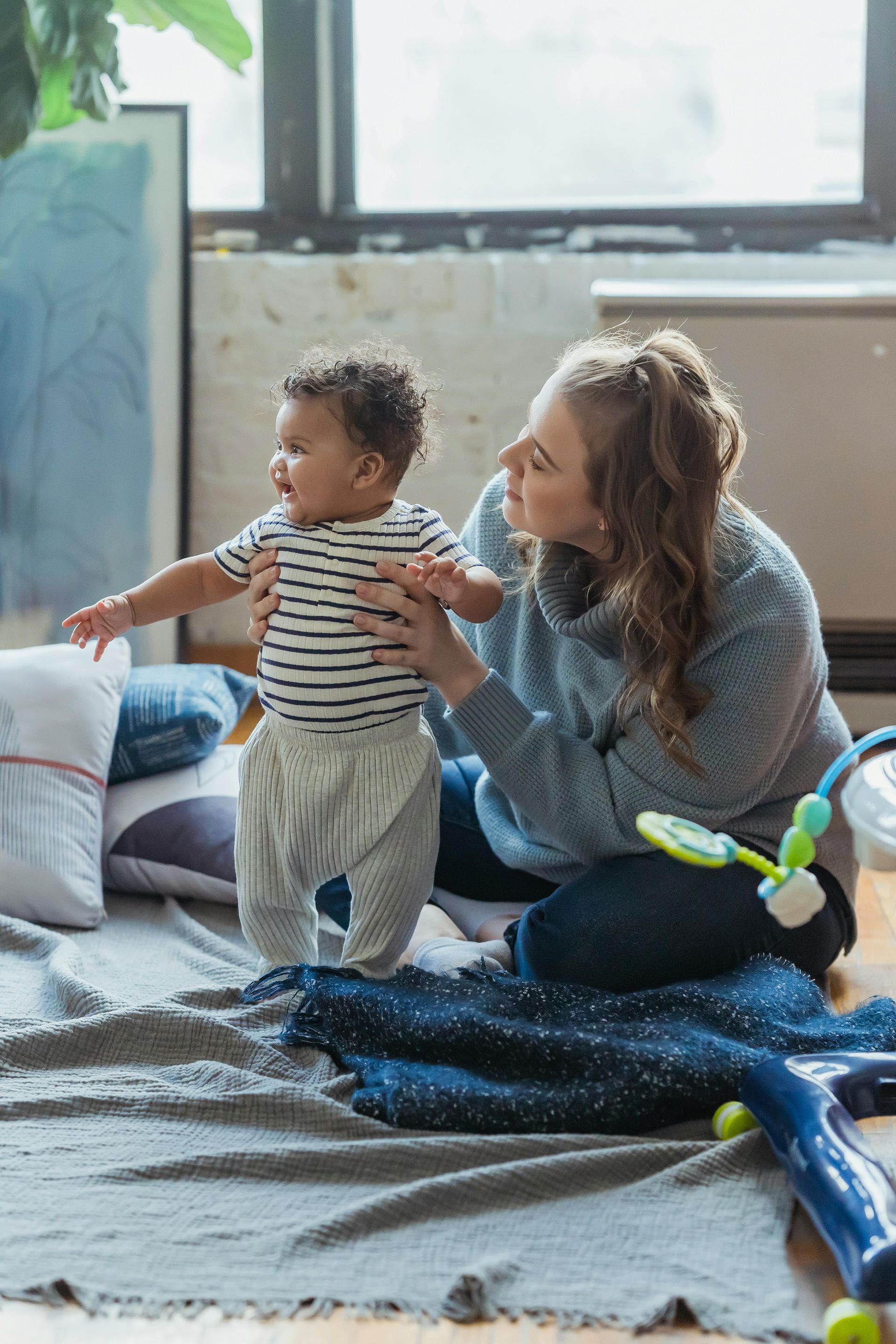 Woman helps baby stand; they are on a rug, near a window.