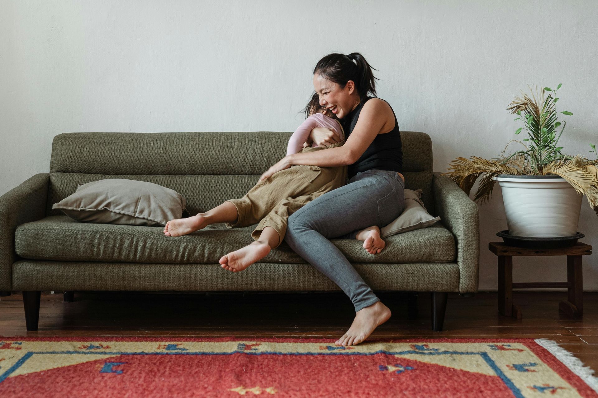 Woman embraces a child on a green couch in a living room; red rug in foreground.