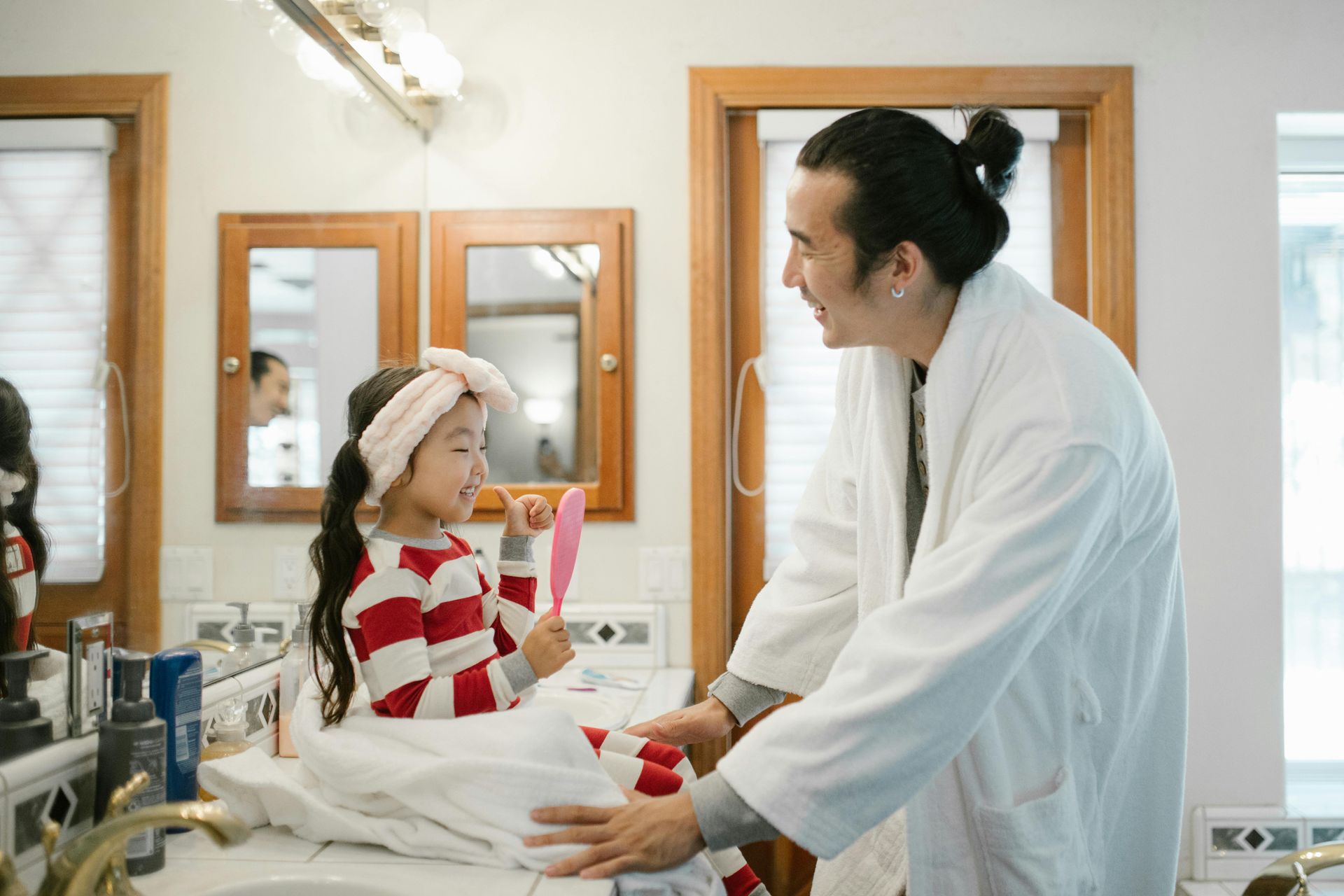A child in pajamas smiles while looking in a hand mirror. An adult with a bun looks on. In a bathroom.