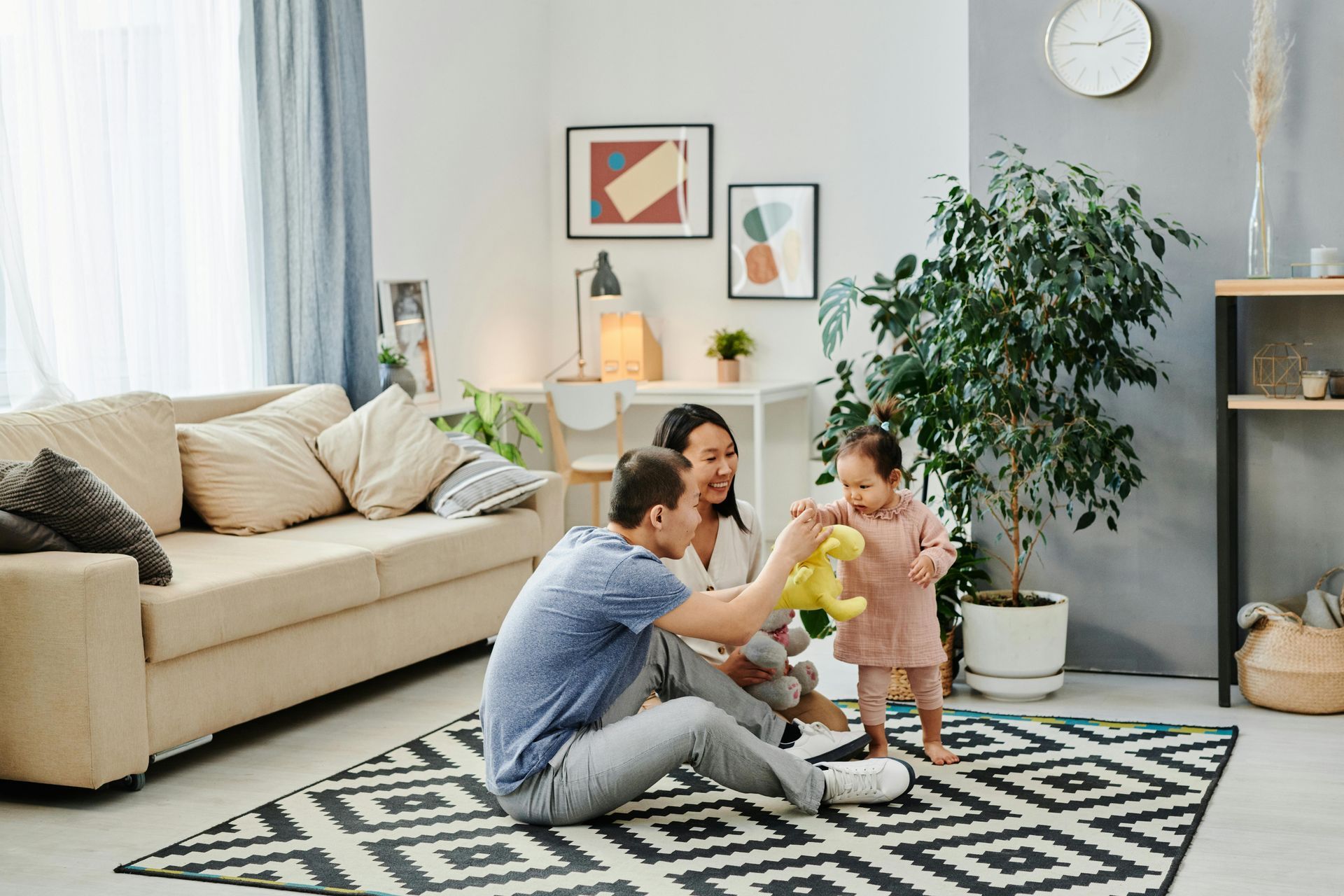 Family plays with toddler in living room. Neutral colored sofa, patterned rug, plants.