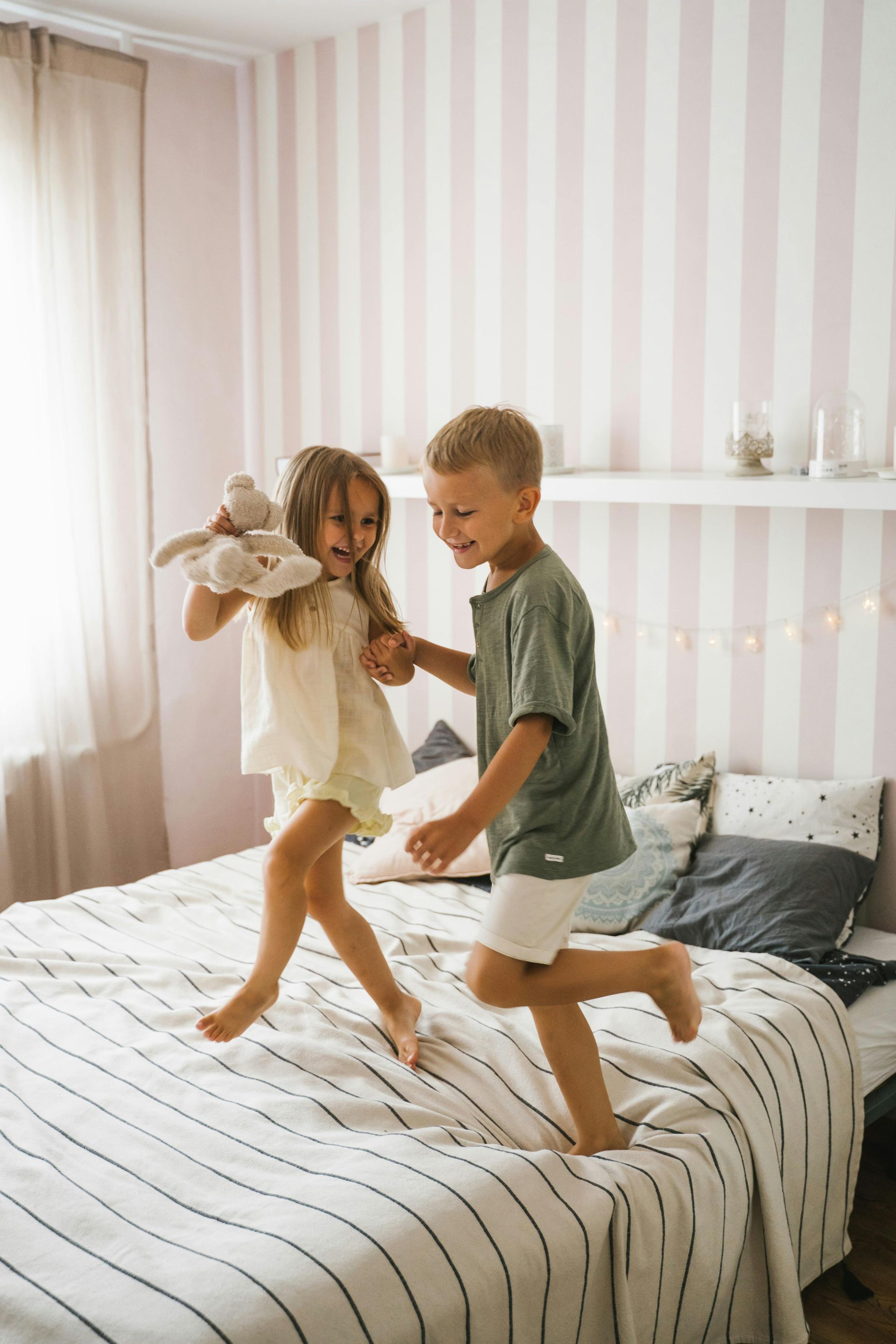 Two children jumping and laughing on a bed with striped bedding; striped pink wall in the background.