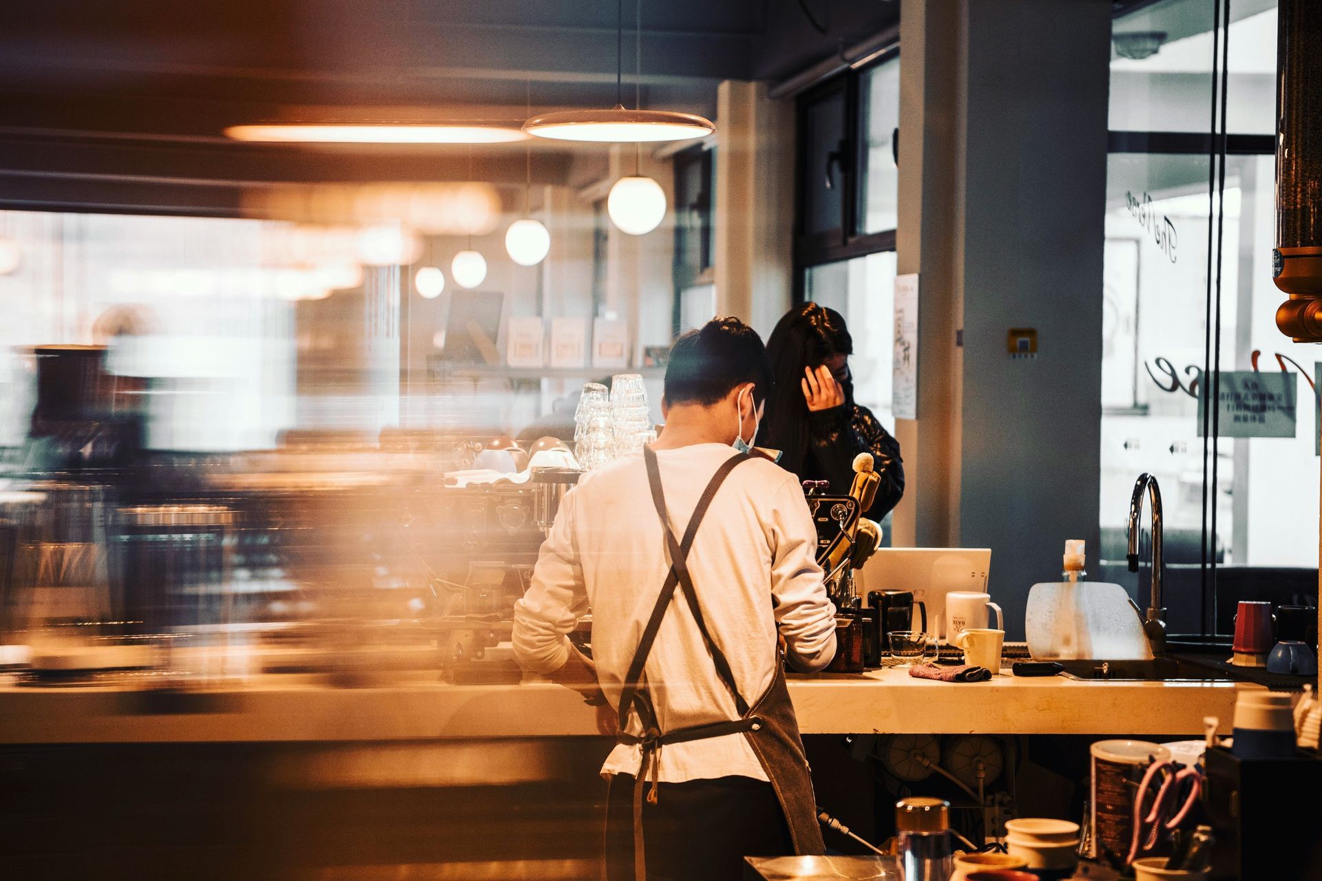Barista wearing an apron behind a counter, blurred motion. Another person talks near the counter. Interior setting.
