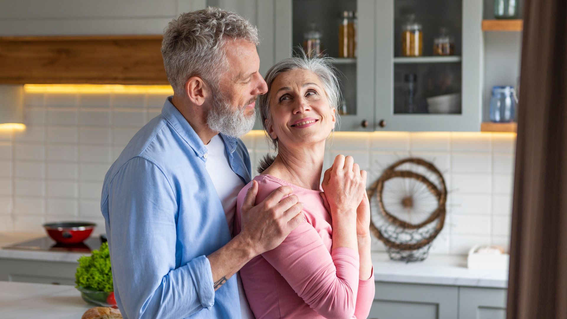 Older couple embracing in a kitchen; man in blue shirt kisses woman's head, both smiling.