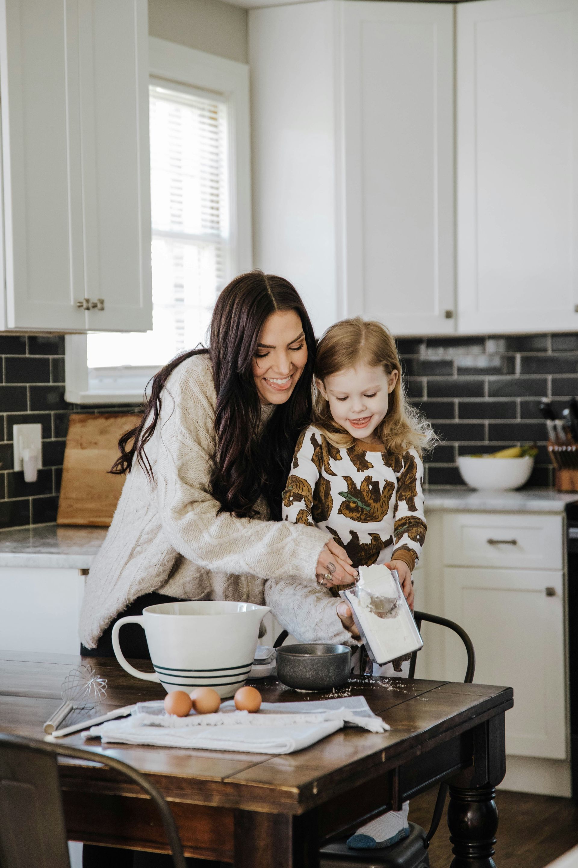 Woman and child baking in a kitchen, pouring ingredients. Child smiles, woman looks on.