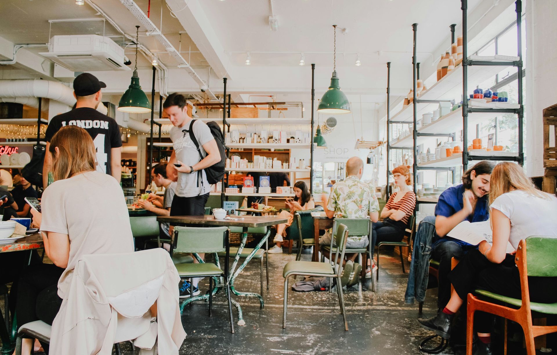 People in a busy cafe, some seated at tables, others standing, with green chairs and shelves.