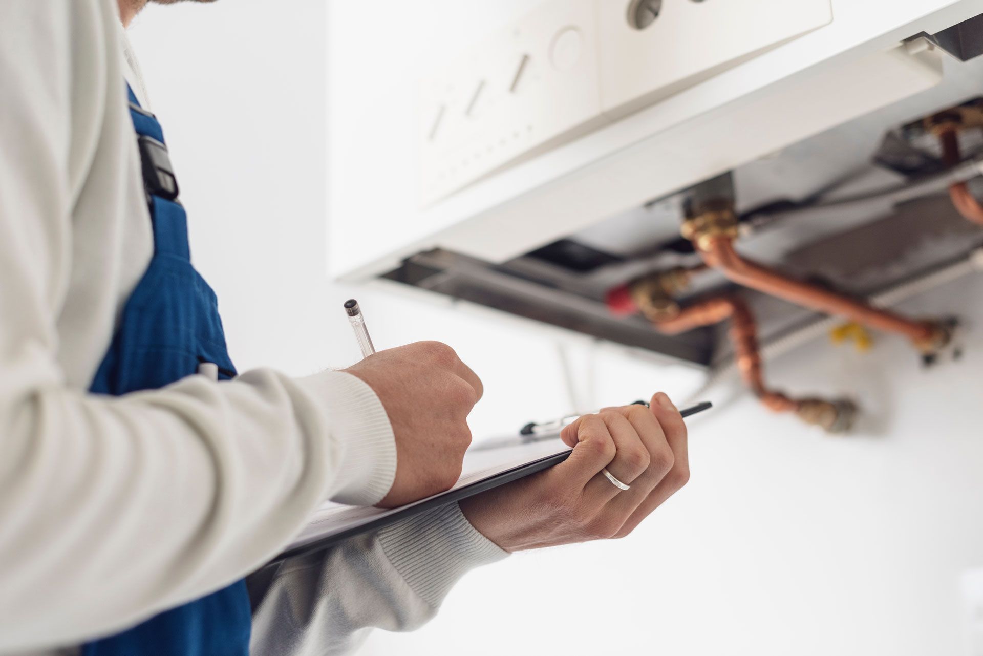 A technician in blue overalls inspects a boiler, writing on a clipboard.