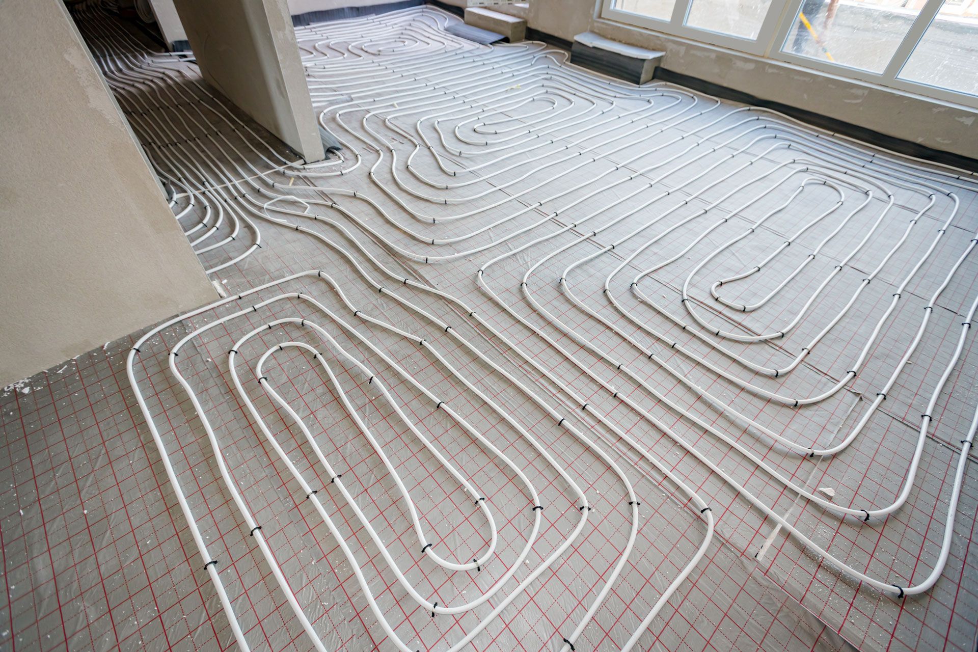 White pipes arranged in a coil pattern across a floor, likely for radiant floor heating, in a construction setting.