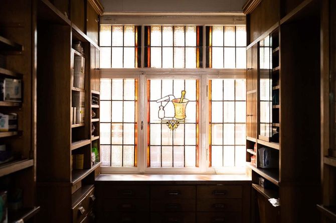 Ein kleiner Apothekenraum mit Holzregalen auf beiden Seiten, der auf ein großes Fenster mit einem Mosaik eines Mörsers und Stößels.