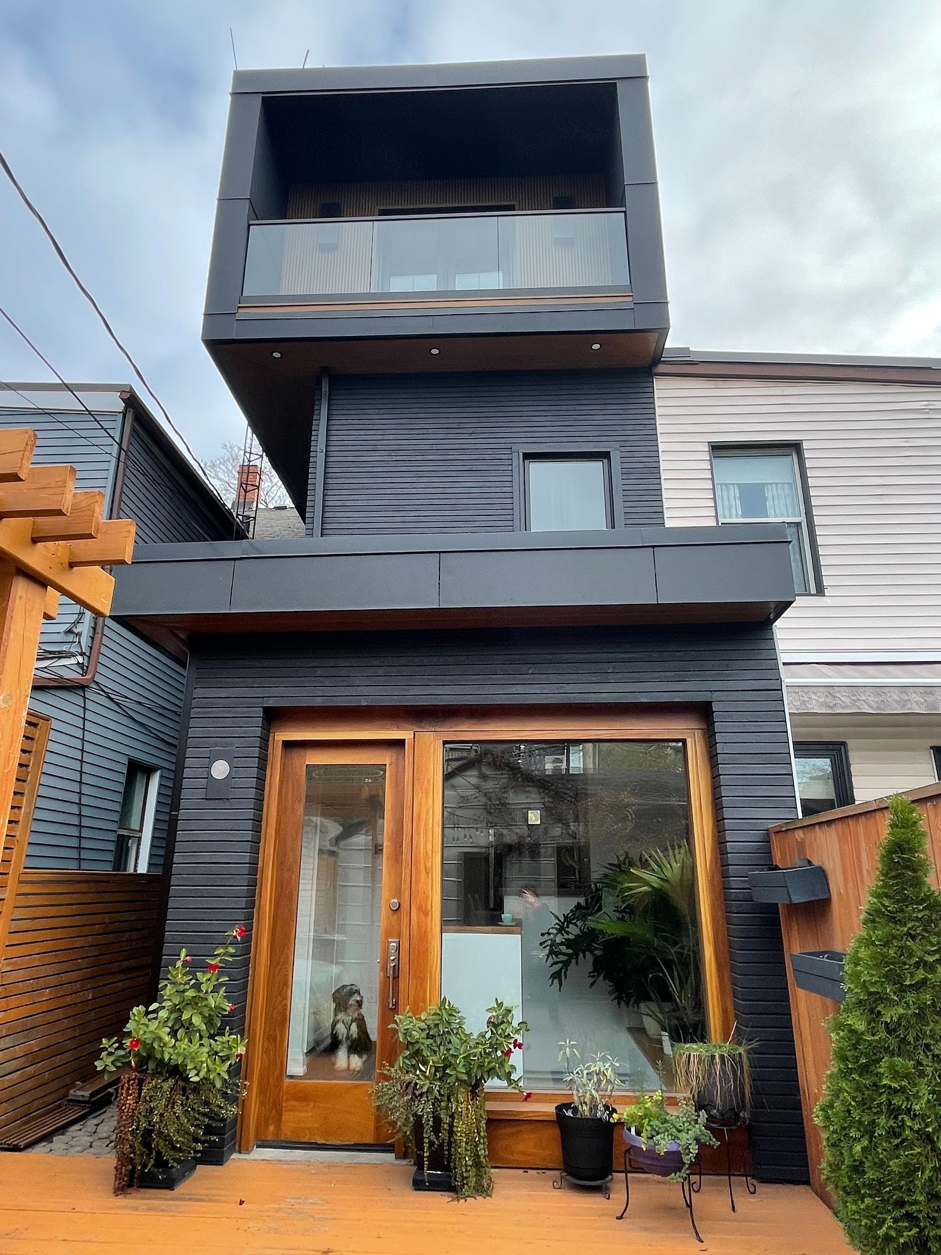 A black and wooden house with a balcony and plants in front of it.