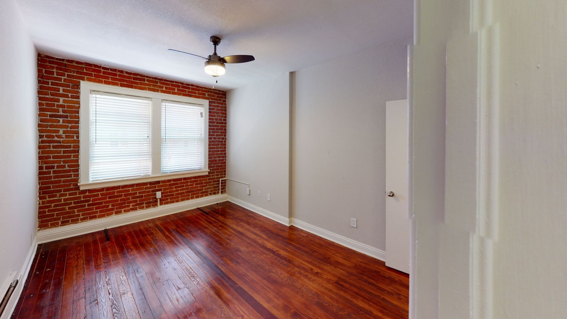 Empty room with hardwood floors, exposed brick wall, window with covering, and ceiling fan.