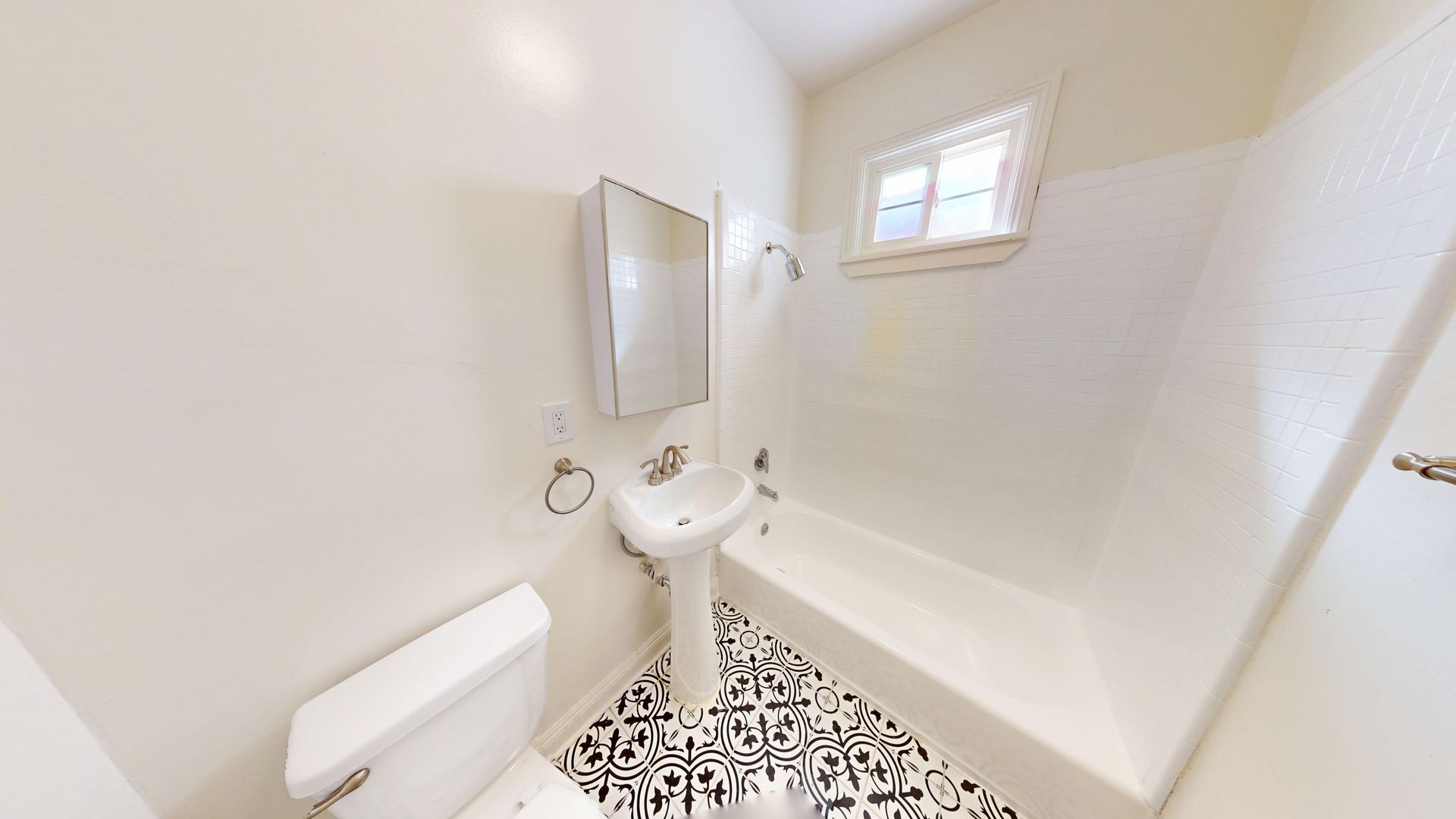 Bathroom with toilet, pedestal sink, bathtub, and patterned floor. White walls, small window.