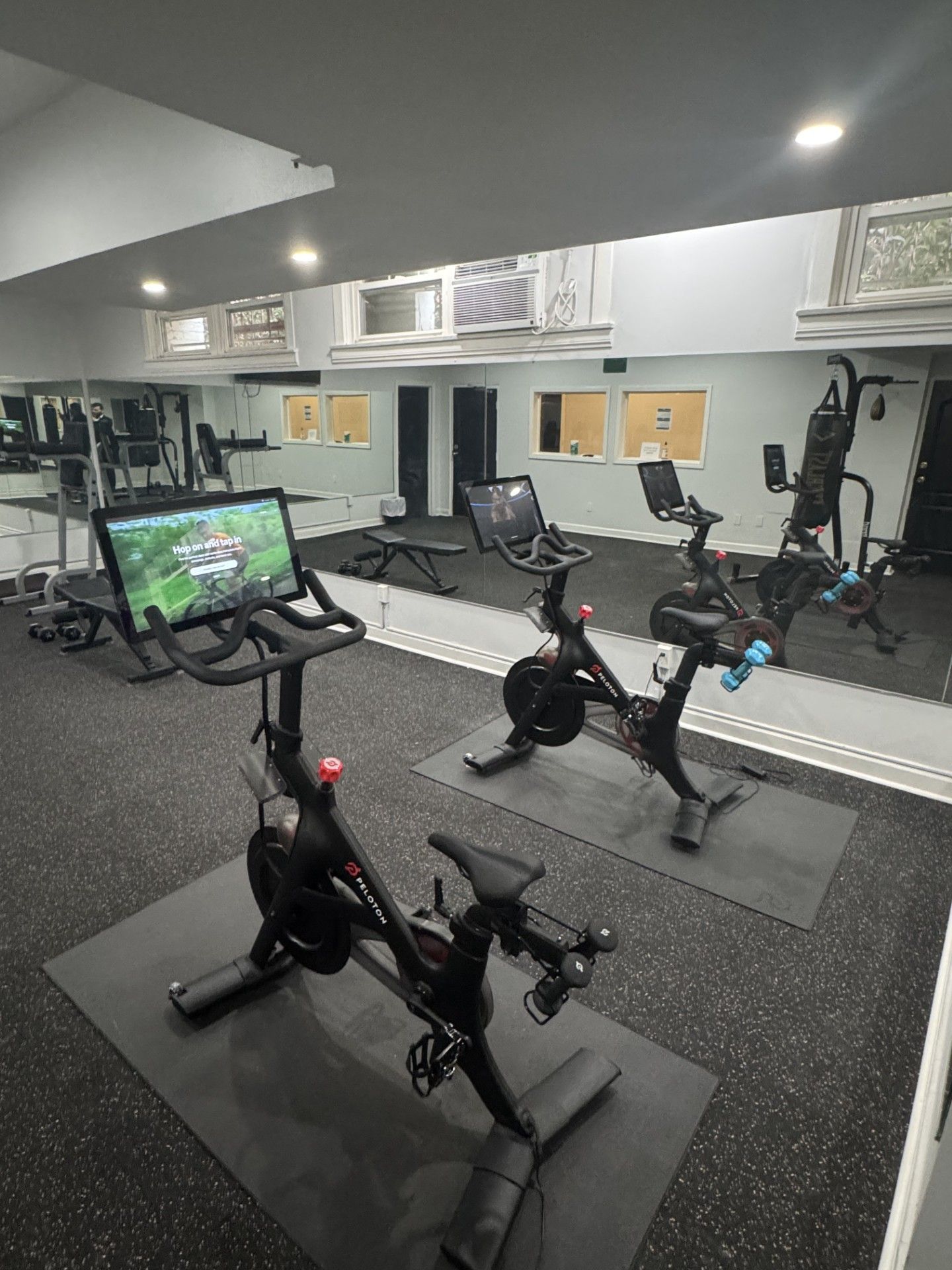 Indoor gym with two stationary bikes, weights, and mirrors. Black exercise mats and gray walls.