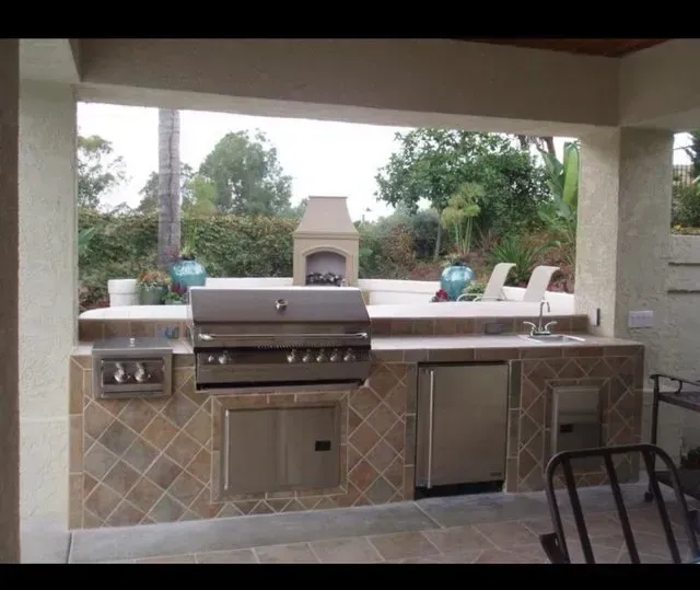 Outdoor kitchen with grill, sink, refrigerator, and fireplace, under a covered patio.