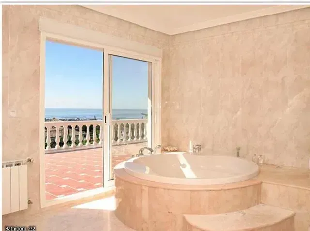 Bathroom with a large, round jacuzzi, large window overlooking a balcony and the sea.