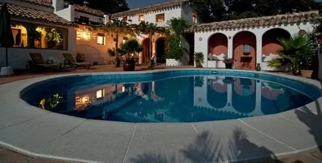 Swimming pool in front of a Spanish-style house at dusk. Pool is blue with white trim. House has arches and red accents.