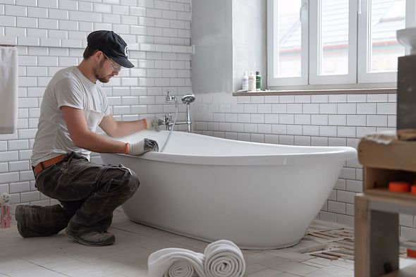 Man installing a white bathtub in a bathroom with white tile walls and a window.