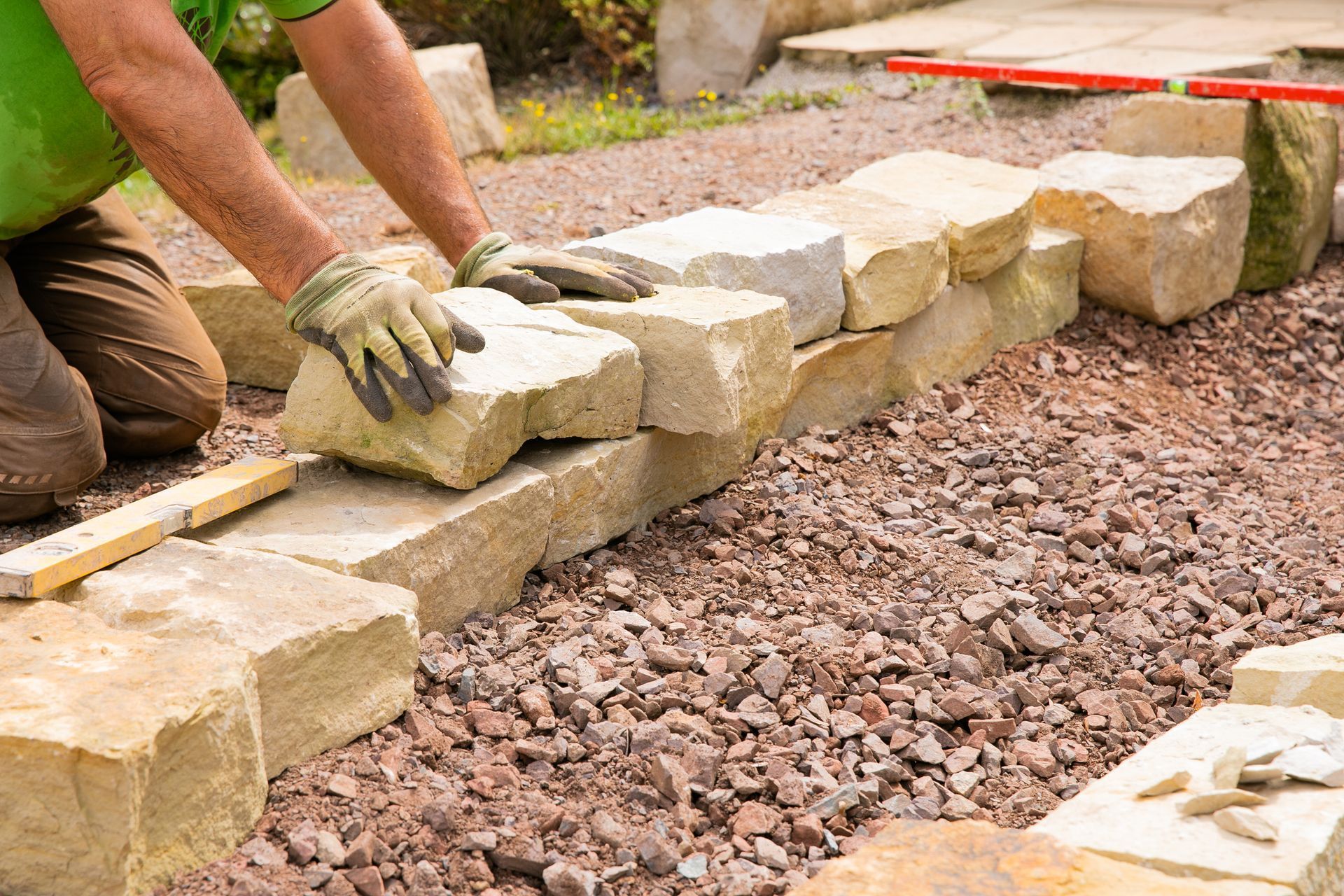 Person wearing gloves building a stone wall in a garden setting, placing a large stone.
