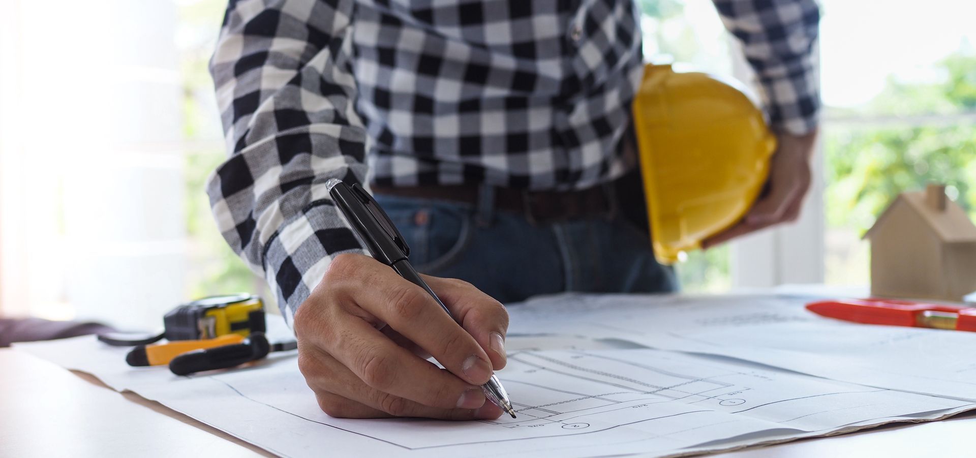 Person in plaid shirt writing on a blueprint, holding a yellow hard hat.