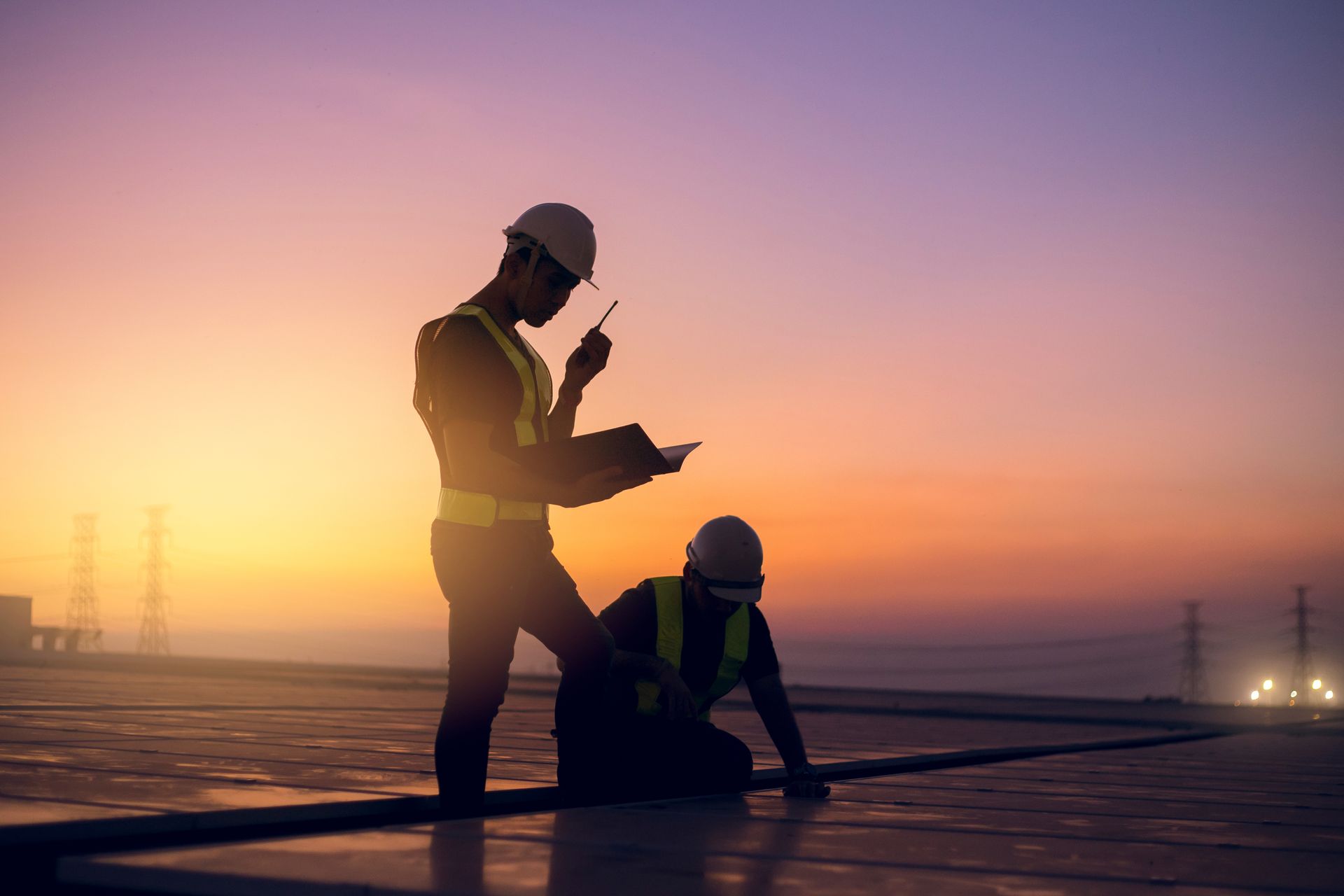 Two construction workers on a rooftop at sunset, one using a radio and the other kneeling.