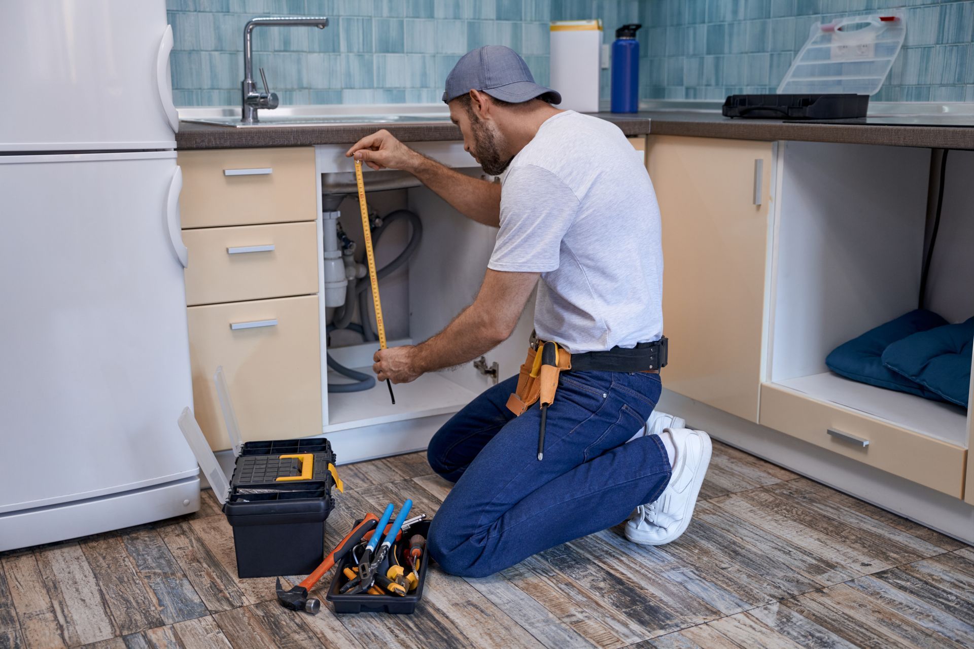 Plumber measuring under a kitchen sink. He is kneeling, wearing a tool belt and hat, with tools nearby.