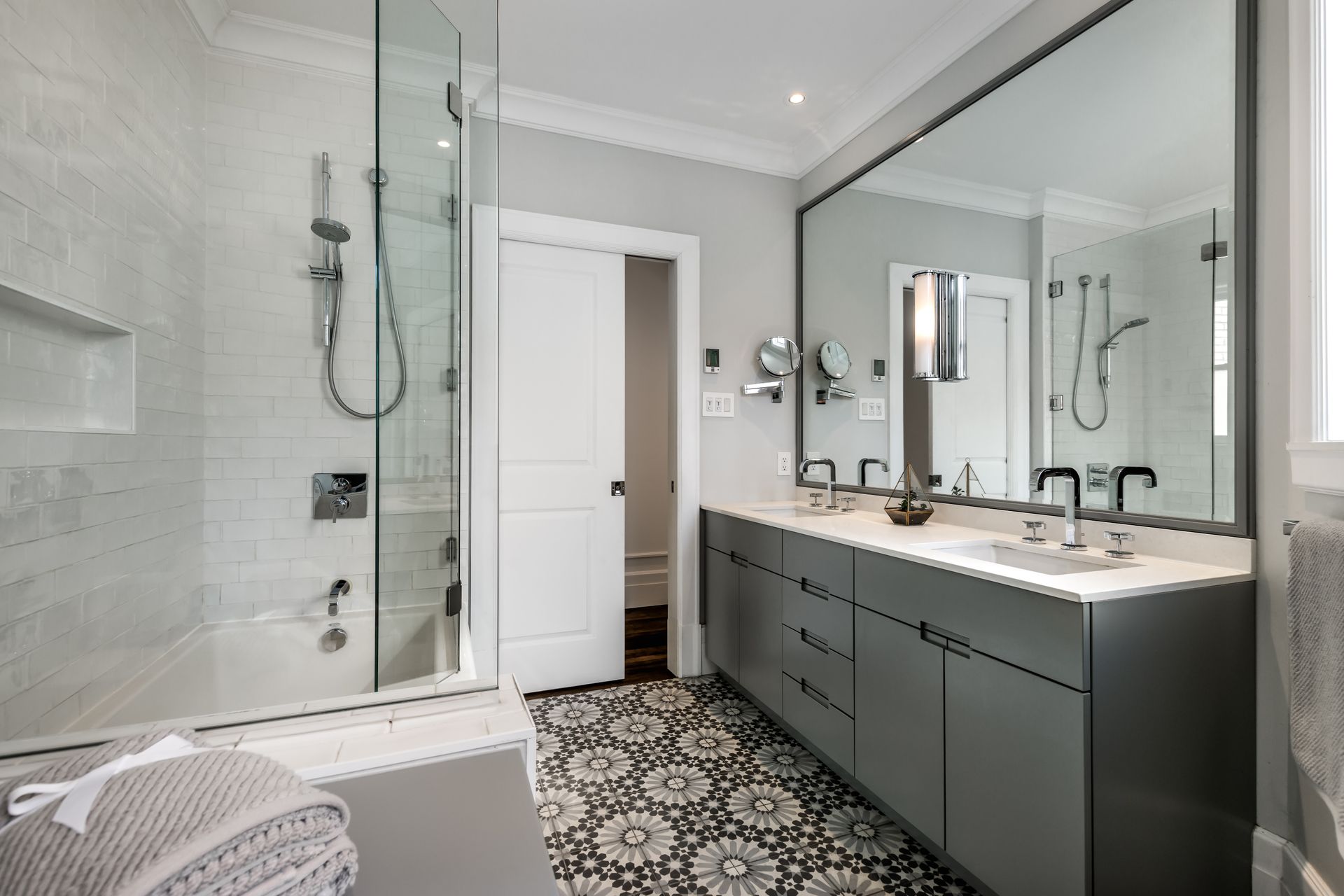 Bathroom with gray vanity, large mirror, glass shower, patterned floor, and white walls.