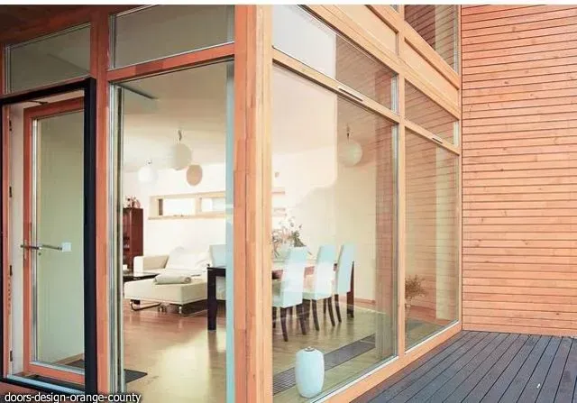 Modern home exterior with large glass windows and door, showcasing interior dining and living areas, and wood siding.