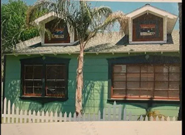 Green house with two dormers and a palm tree in front, behind a white picket fence.