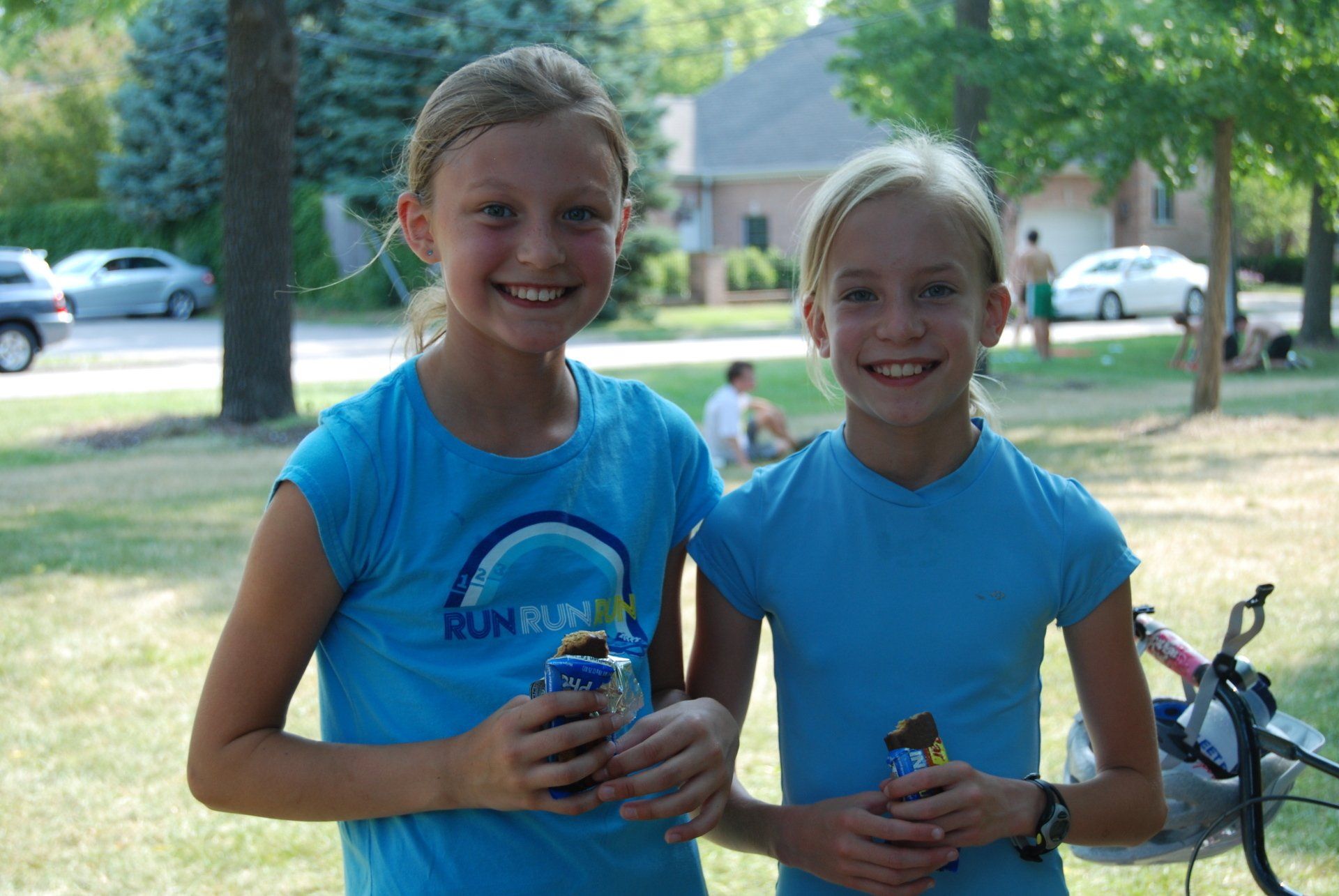 Two young girls are standing next to each other in a park holding snacks.