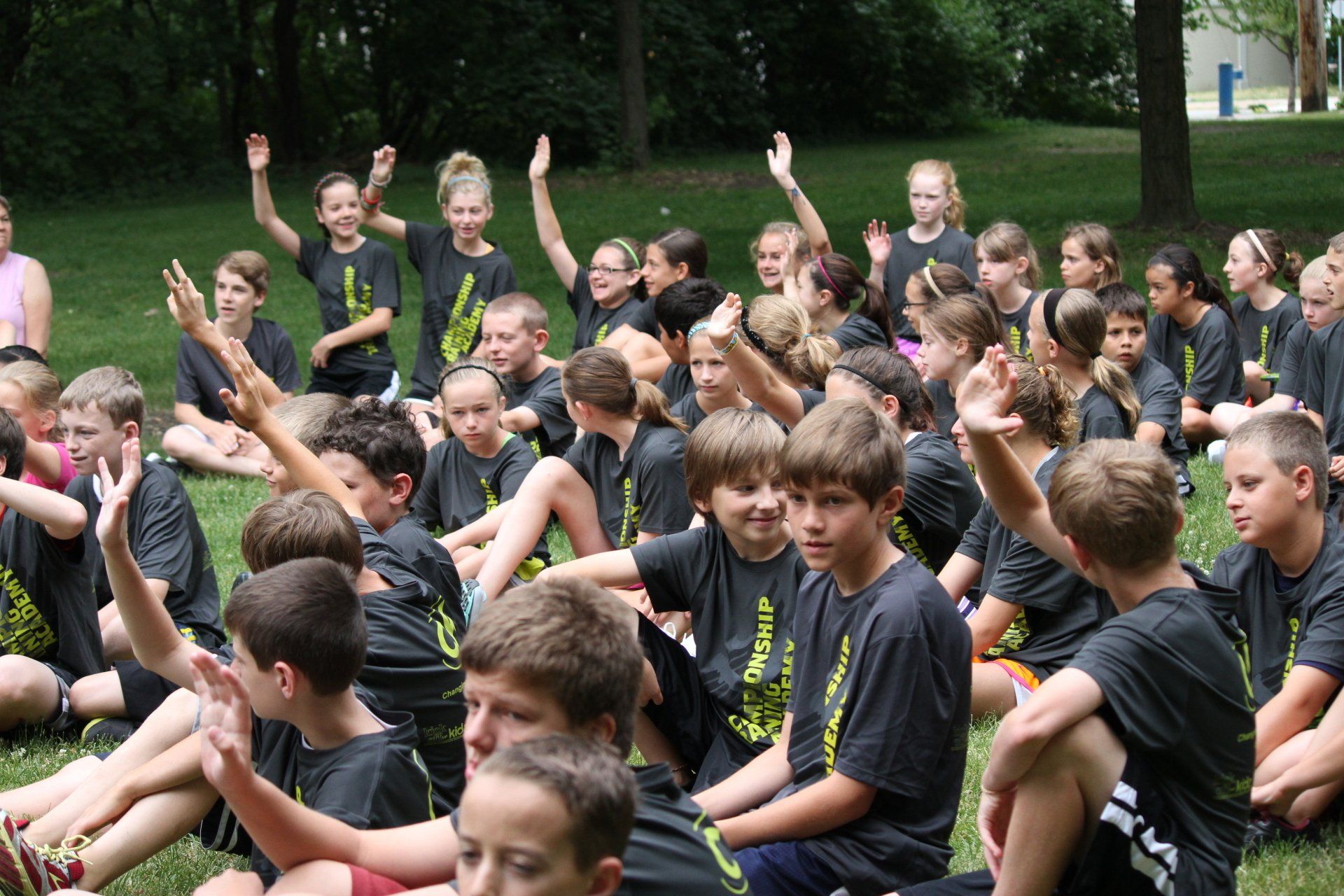 A group of children are sitting in the grass raising their hands