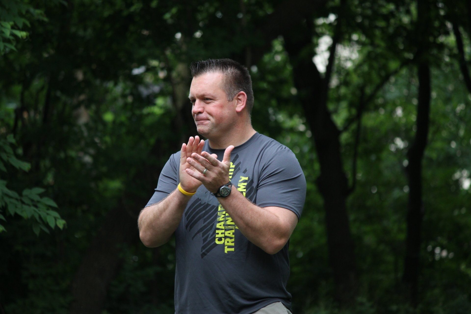 A man in a gray shirt is clapping his hands in the woods.